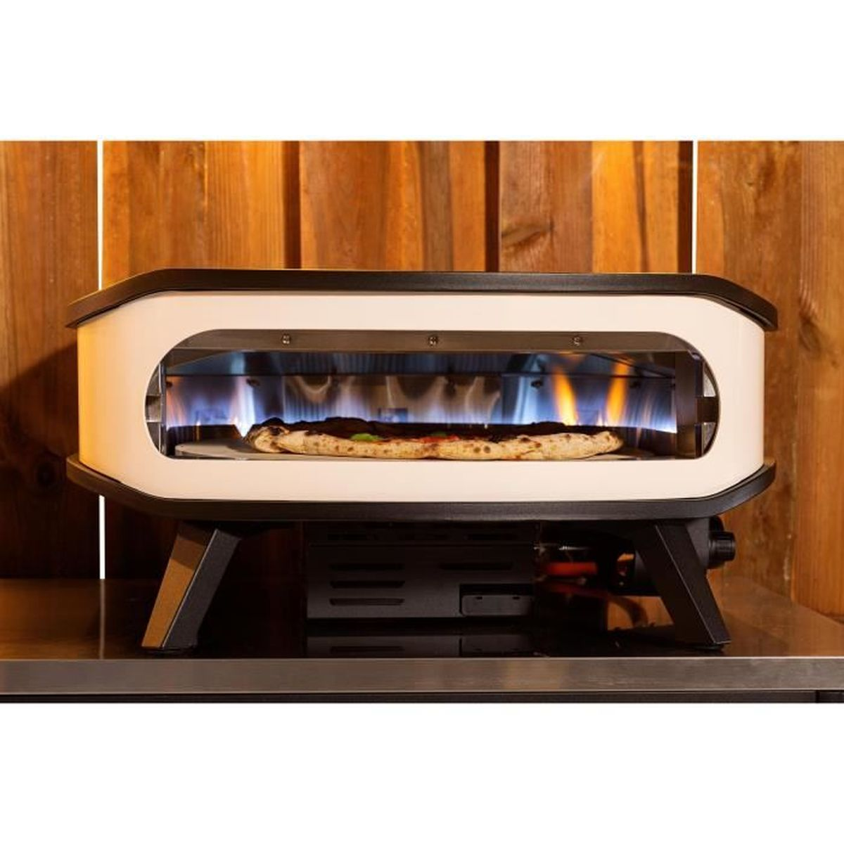 Een pizzaoven met een pizza erin, vlammen zichtbaar. De oven is wit en zwart, op een metalen oppervlak, tegen een houten achtergrond.