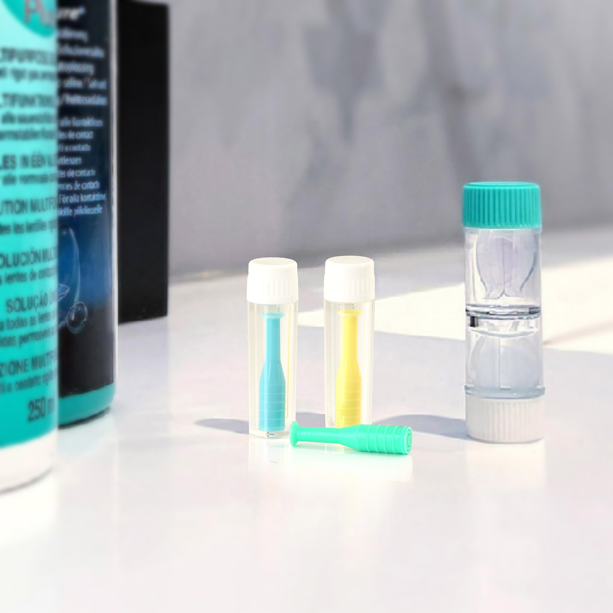 Zubehör für Kontaktlinsen: Behälter, Applikatoren und Reinigungsflaschen. Farben: Weiß, Blau, Gelb und Grün. Weiße Oberfläche.
