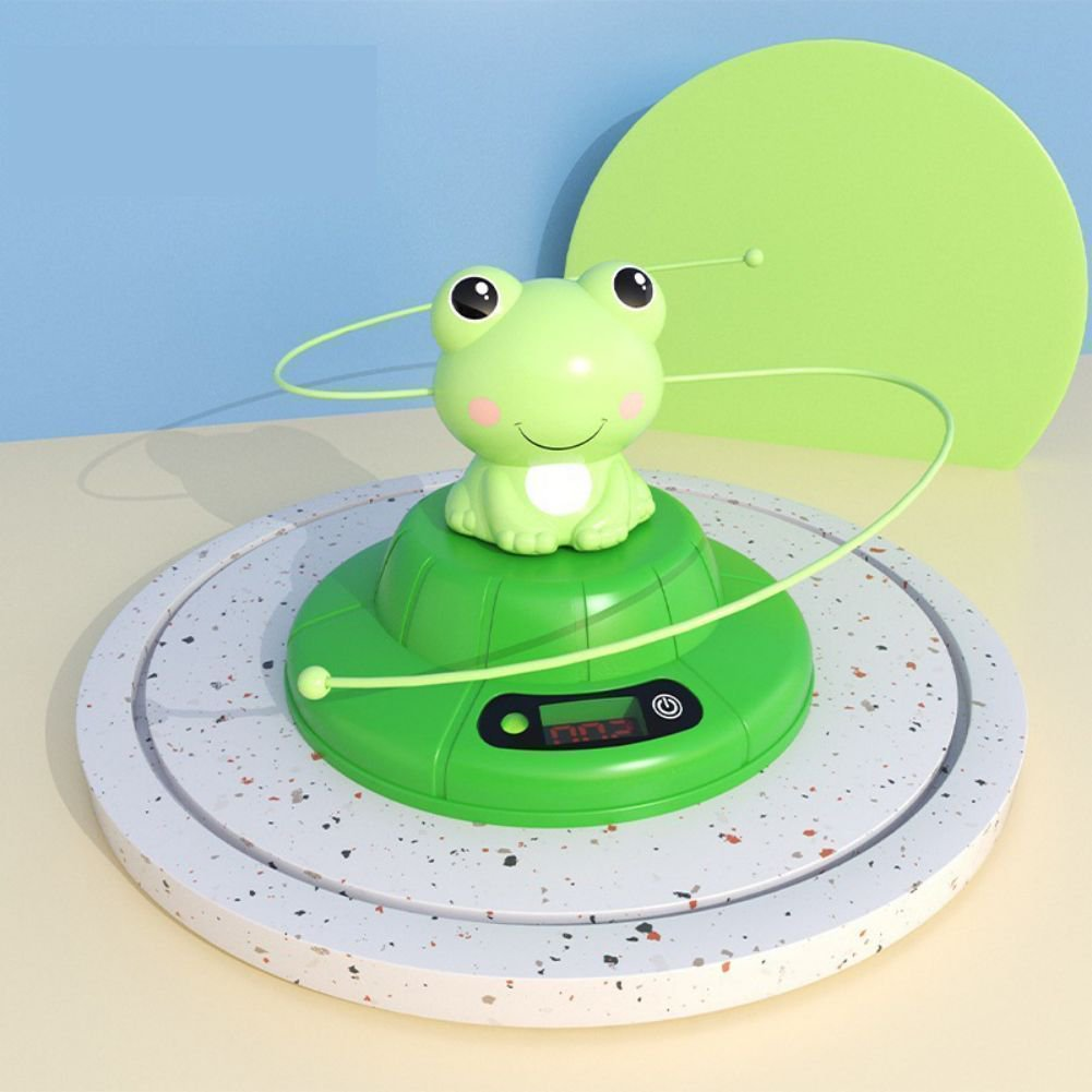 Ein grünes Froschspielzeug auf einer grünen Basis mit digitaler Anzeige. Der Hintergrund hat blaue und grüne Elemente, und das Spielzeug steht auf einer Plattform.