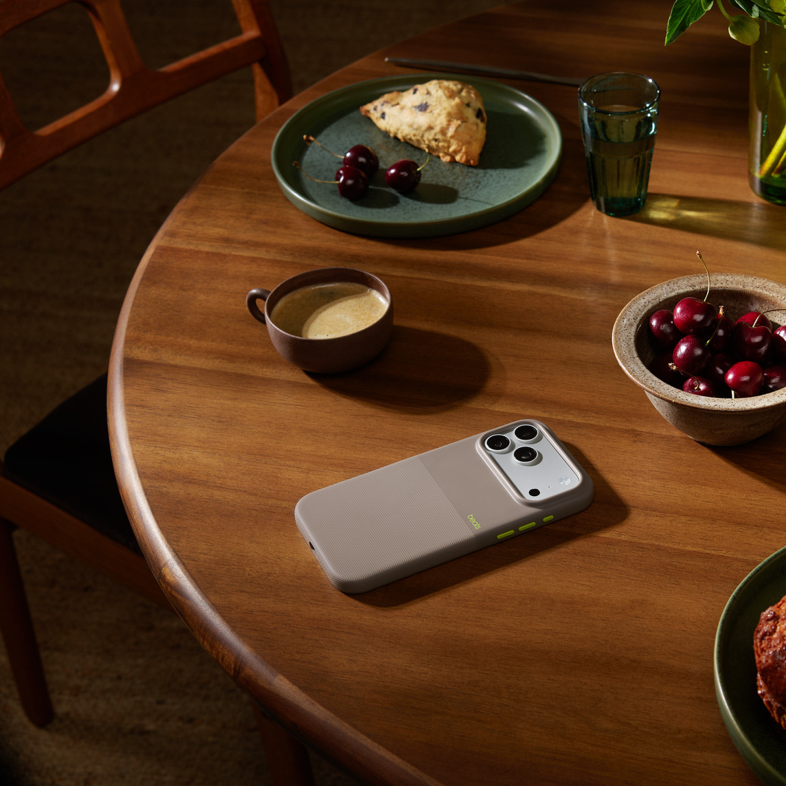 Drewniany stół z telefonem, kawą, wiśniami, ciastkiem i szklanką wody.