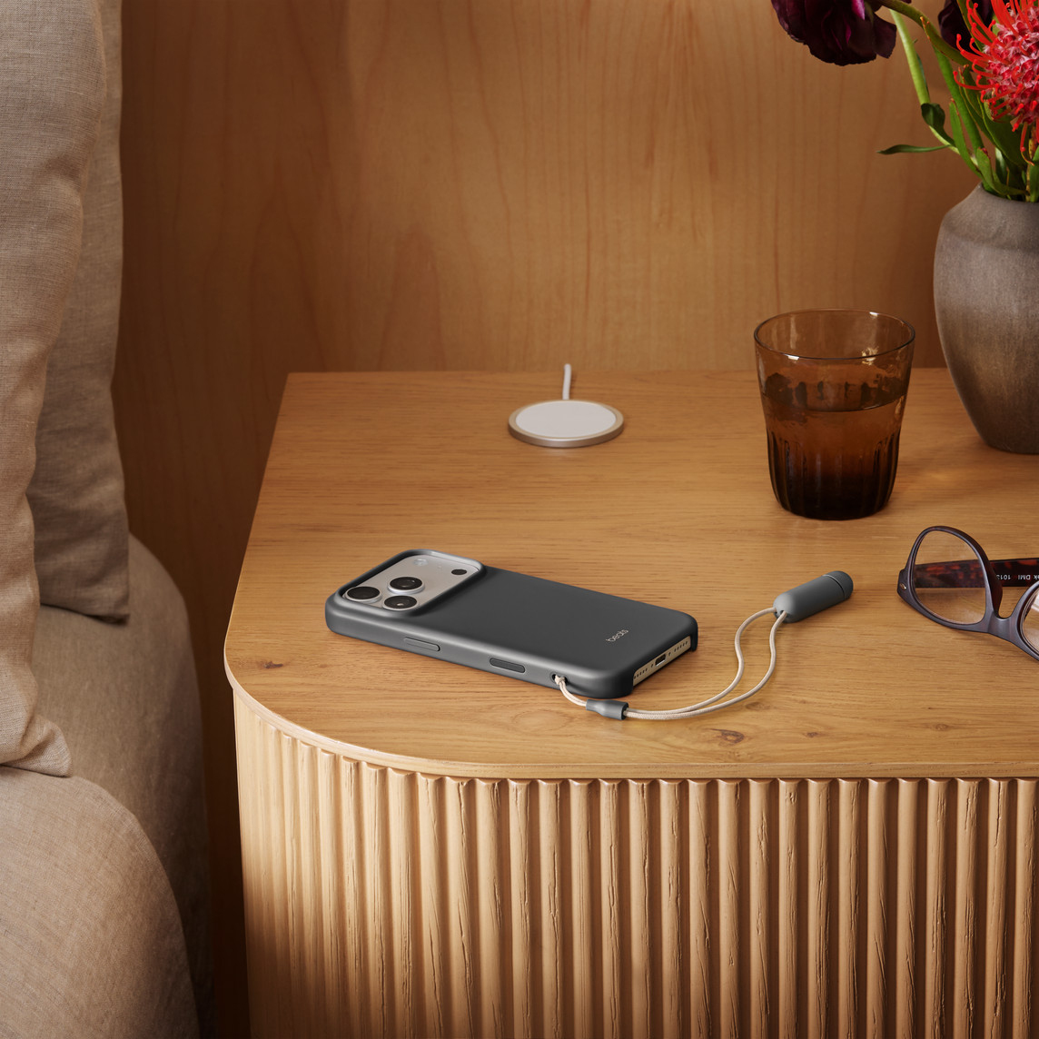 Ciemny telefon z kablem ładującym na drewnianej szafce nocnej. Obecna jest również szklanka wody, kwiaty i okulary.