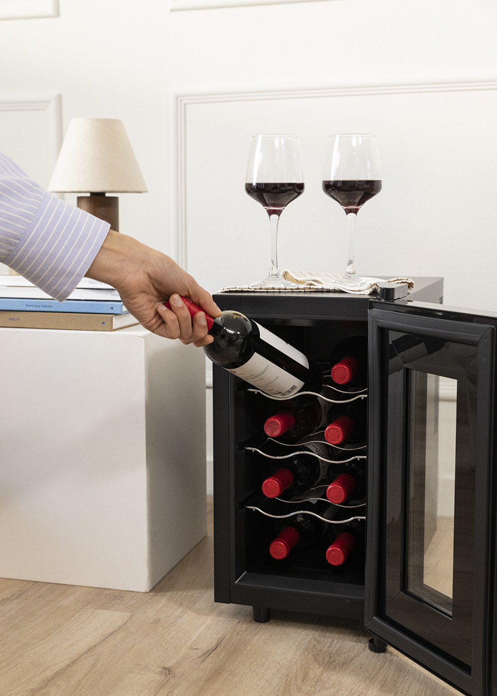 Una mano colocando una botella de vino en un enfriador de vino negro. Dos copas de vino y una lámpara están en el mostrador.