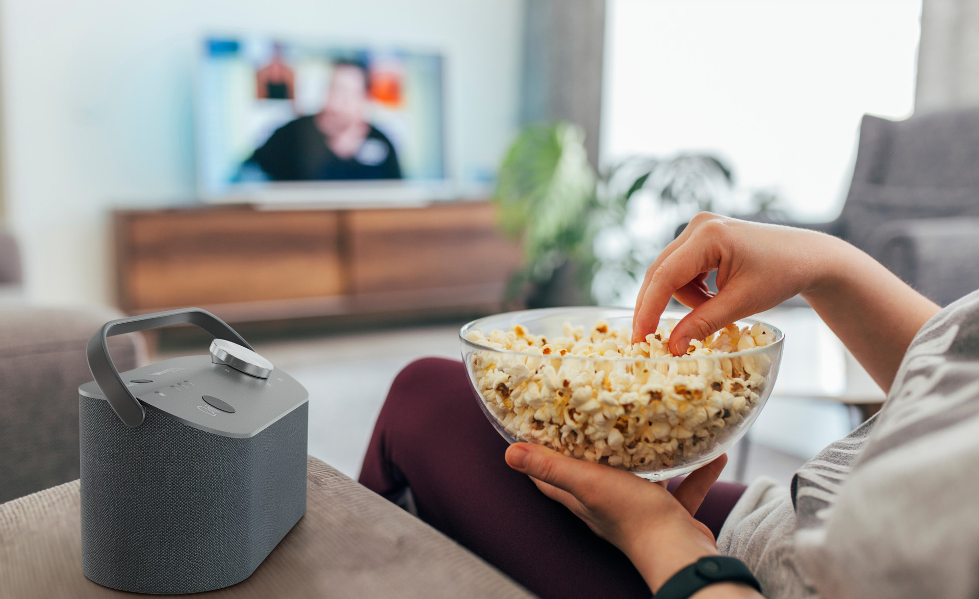Eine Person isst Popcorn aus einer Glasschüssel, während sie fernsieht. Auf dem Tisch steht ein Lautsprecher.
