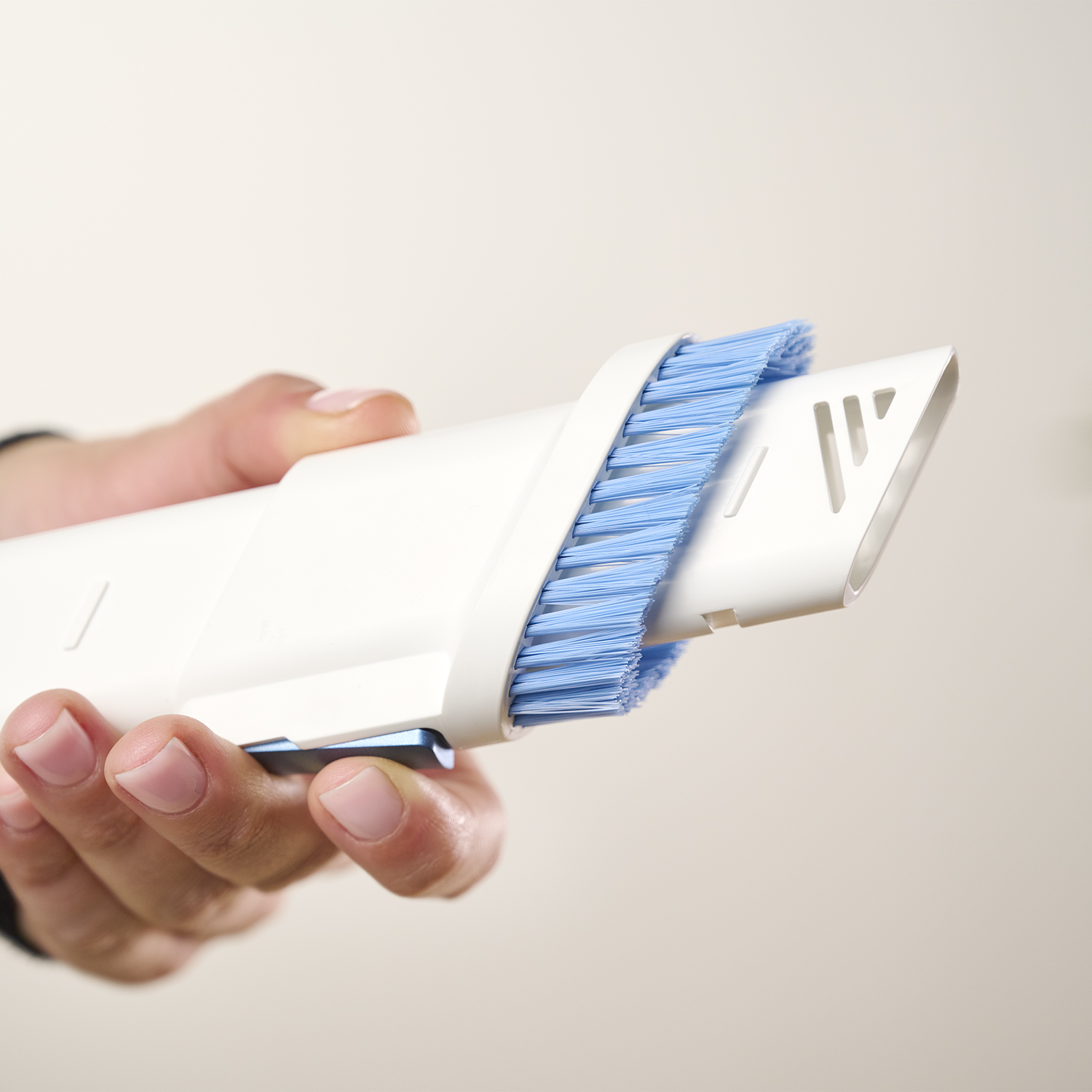 Una mano tiene un accessorio bianco per aspirapolvere con setole blu. Lo sfondo è beige chiaro uniforme.