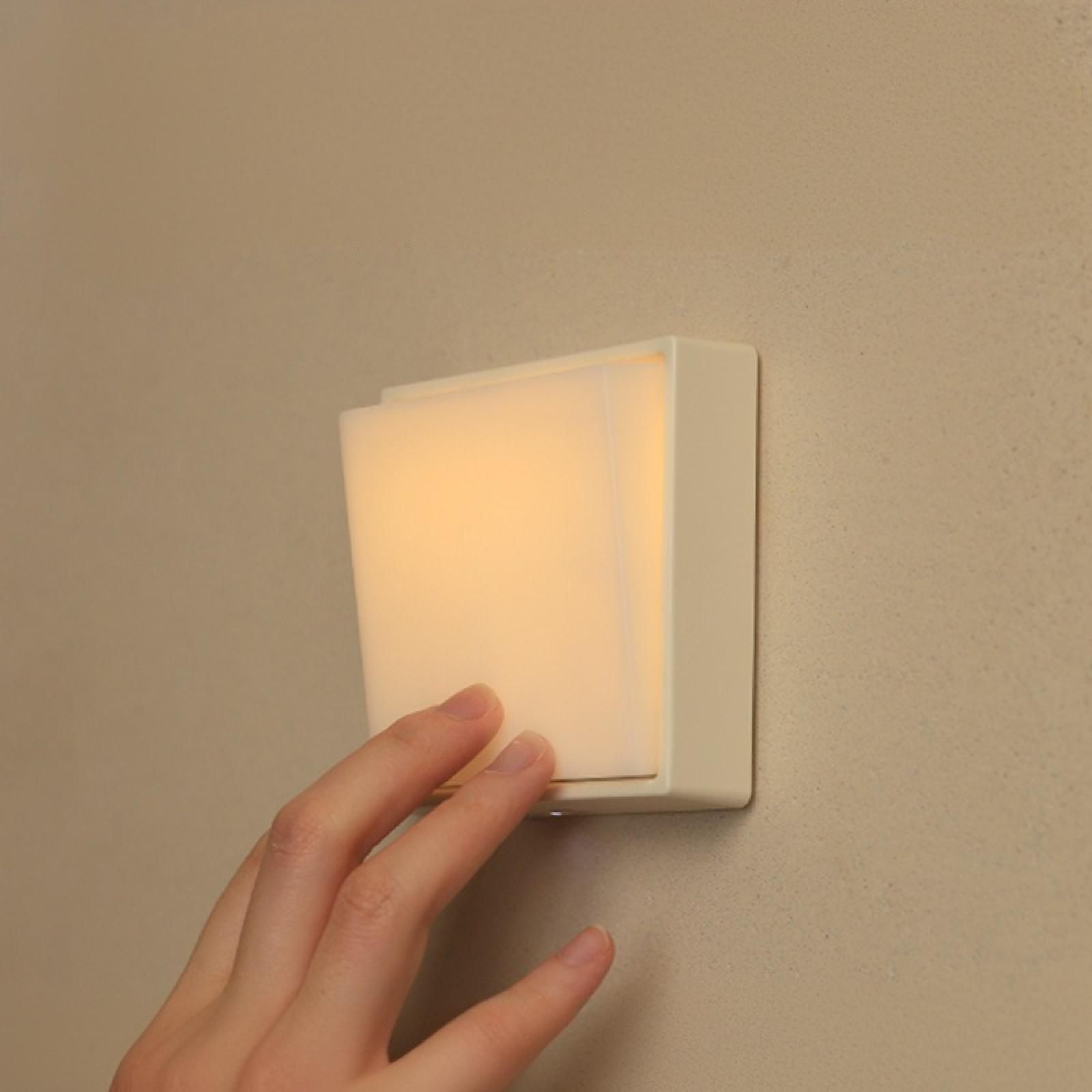 Eine Hand berührt eine quadratische, leuchtende Leuchte an einer beigefarbenen Wand. Das Licht ist weich und warm.