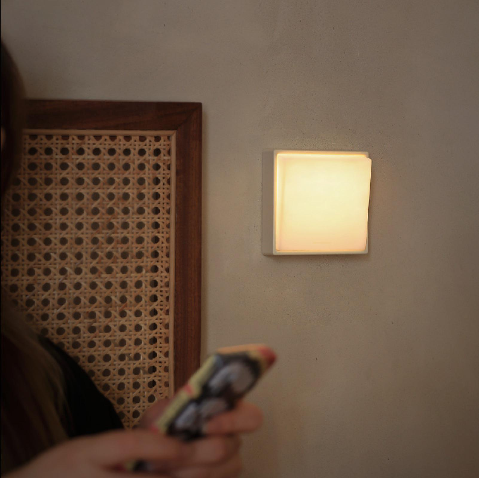 Eine leuchtende quadratische Lampe an der Wand. Eine Person hält ein Telefon. Holzrahmen und Webmuster.