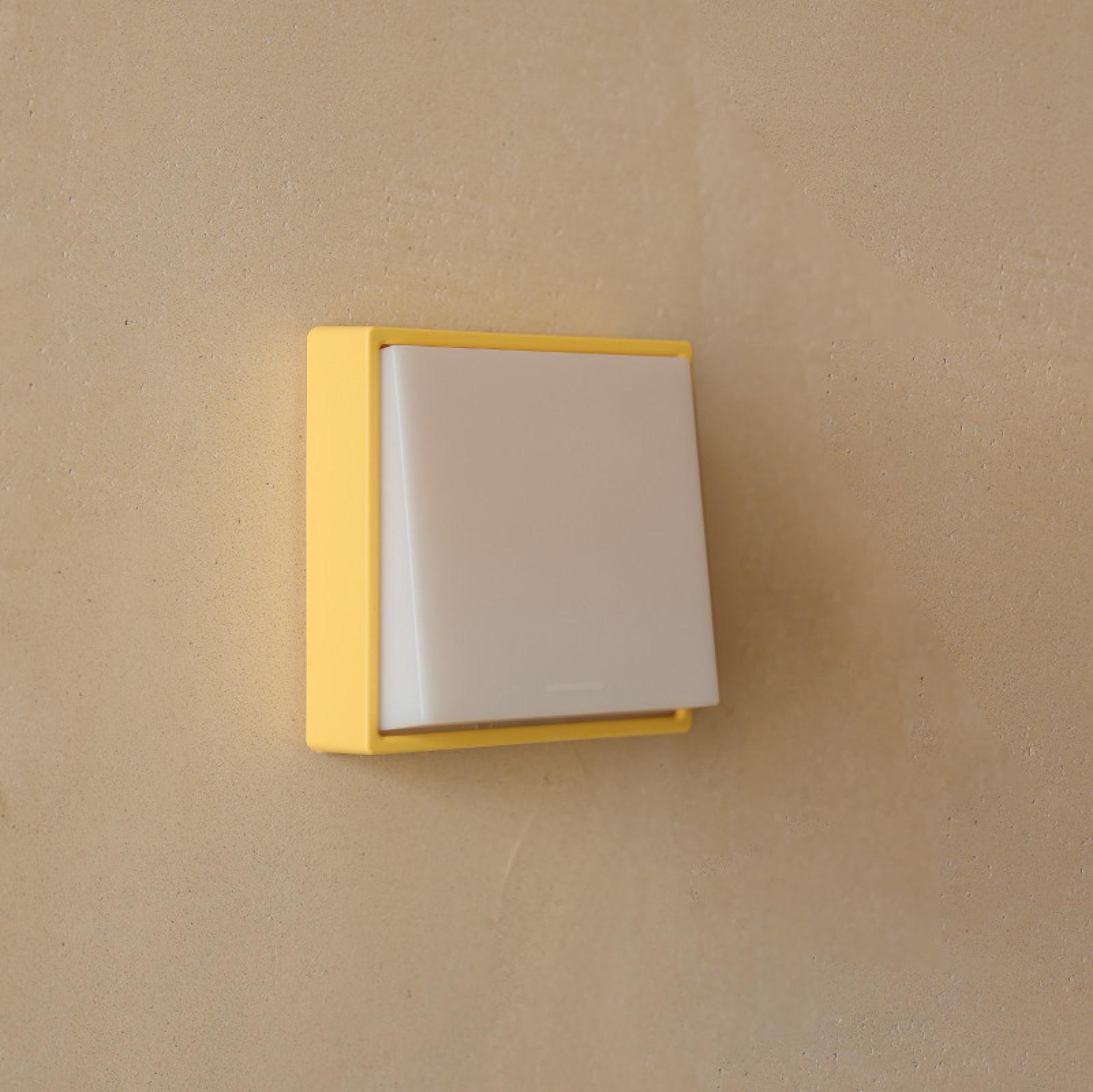 Ein gelb gerahmter Lichtschalter mit weißem, rechteckigem Knopf ist an einer beigefarbenen Wand befestigt.