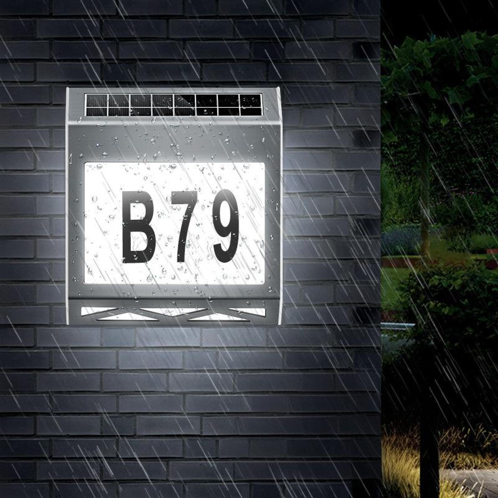 Ein beleuchtetes Hausnummernschild mit 'B79' und Regentropfen. Es ist an einer Ziegelmauer befestigt. Rechts ein Baum.