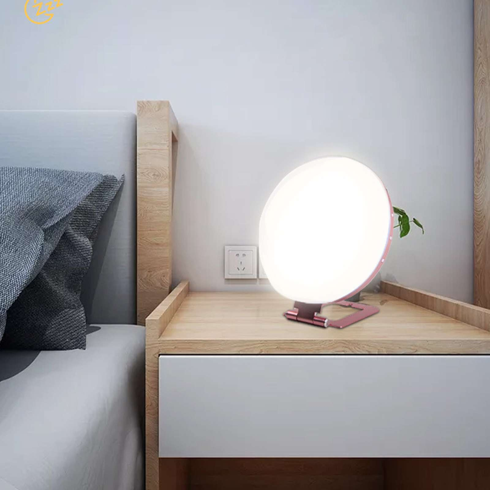 Eine runde Lampe leuchtet auf einem Nachttisch neben einem Bett. Das Licht der Lampe ist hell. Neben der Lampe steht eine Pflanze.