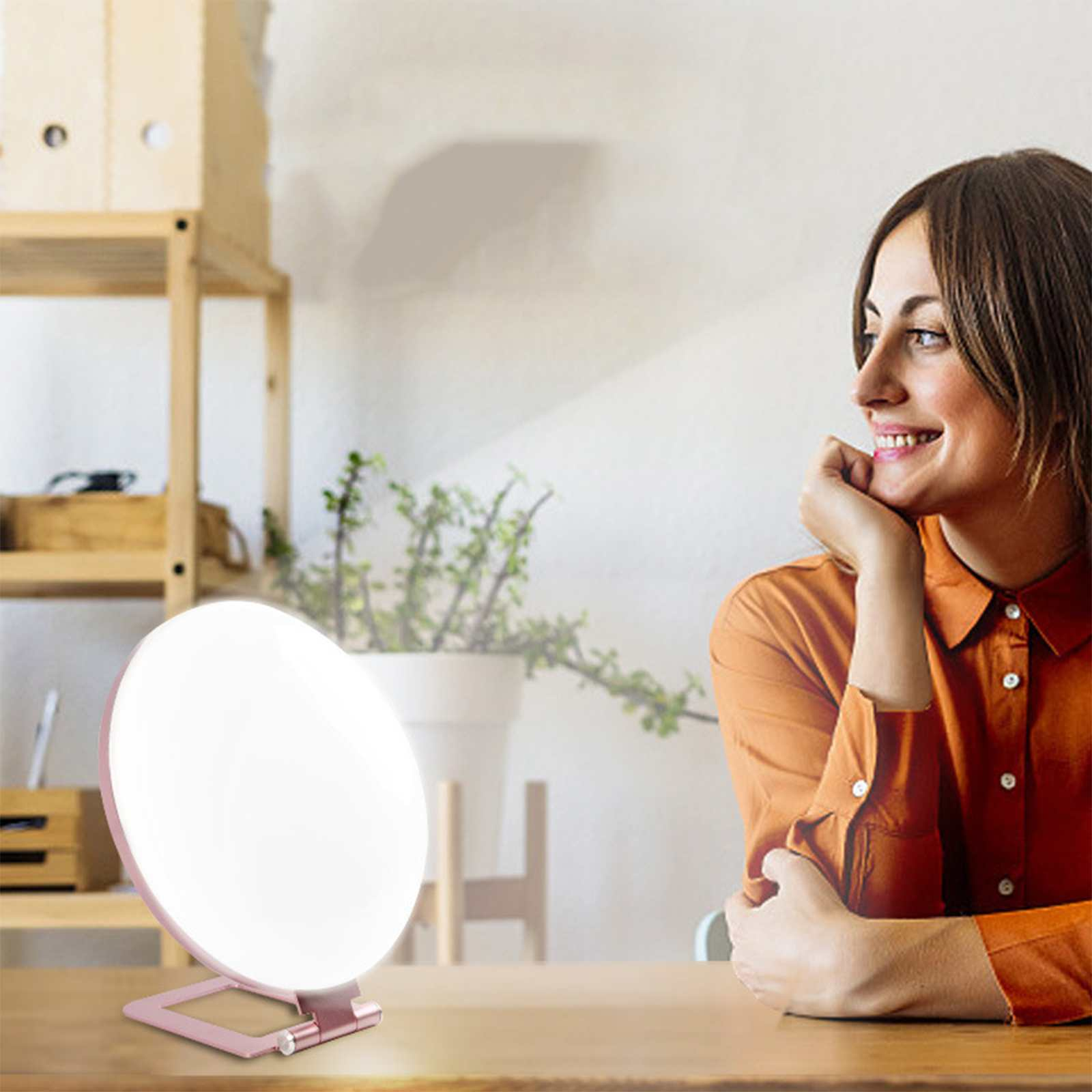 Eine Frau lächelt in der Nähe einer Lichttherapielampe. Die Lampe steht auf einem Holztisch. Im Hintergrund sind eine Pflanze und Regale.