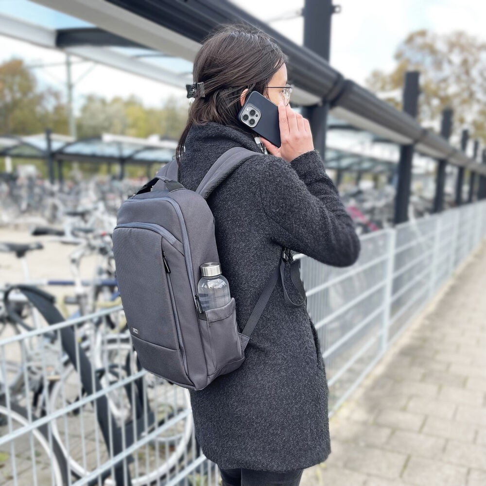 Een persoon met een bril en een rugzak belt. Ze staan in de buurt van fietsenrekken. Een waterfles zit in de rugzak.