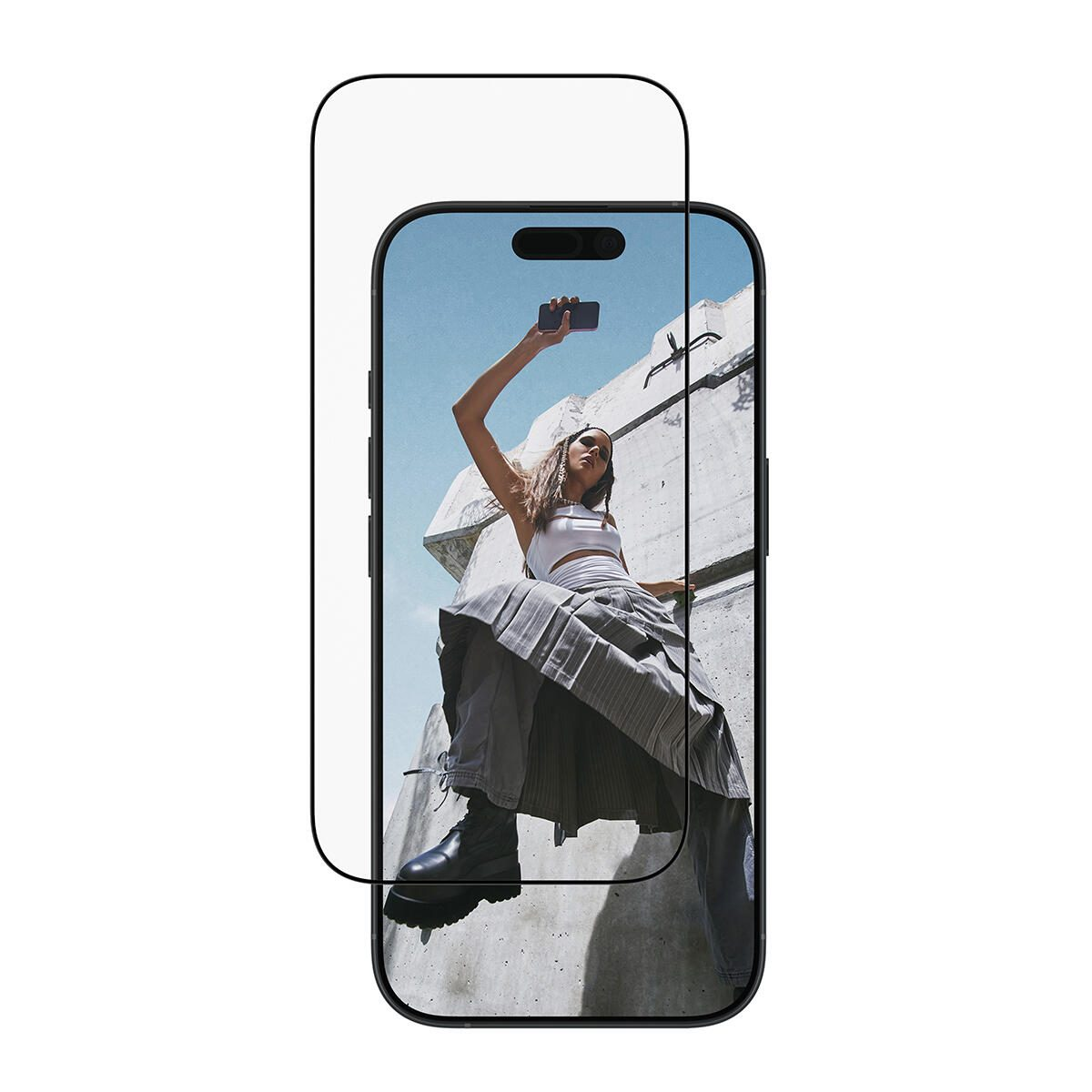 Bir telefon, selfie çeken bir kadını gösteriyor. Beton bir yapıda, beyaz bir üst ve gri etek giyiyor, telefonu tutuyor.
