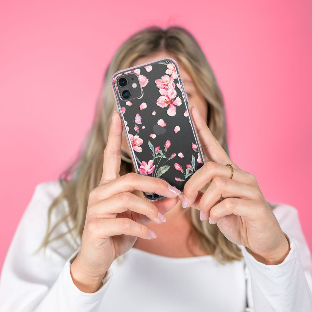 Una donna tiene in mano una custodia per telefono con un design floreale rosa. Sfondo rosa. Indossa una maglia bianca e un anello d'oro.