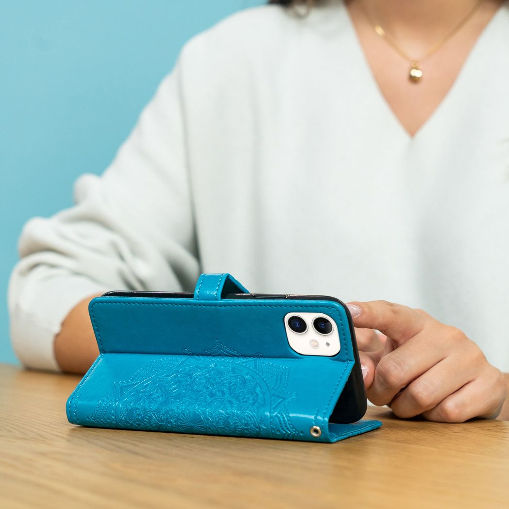 Una persona tiene in mano un telefono in una custodia turchese. Il telefono è su un tavolo di legno. Lo sfondo è un muro blu.