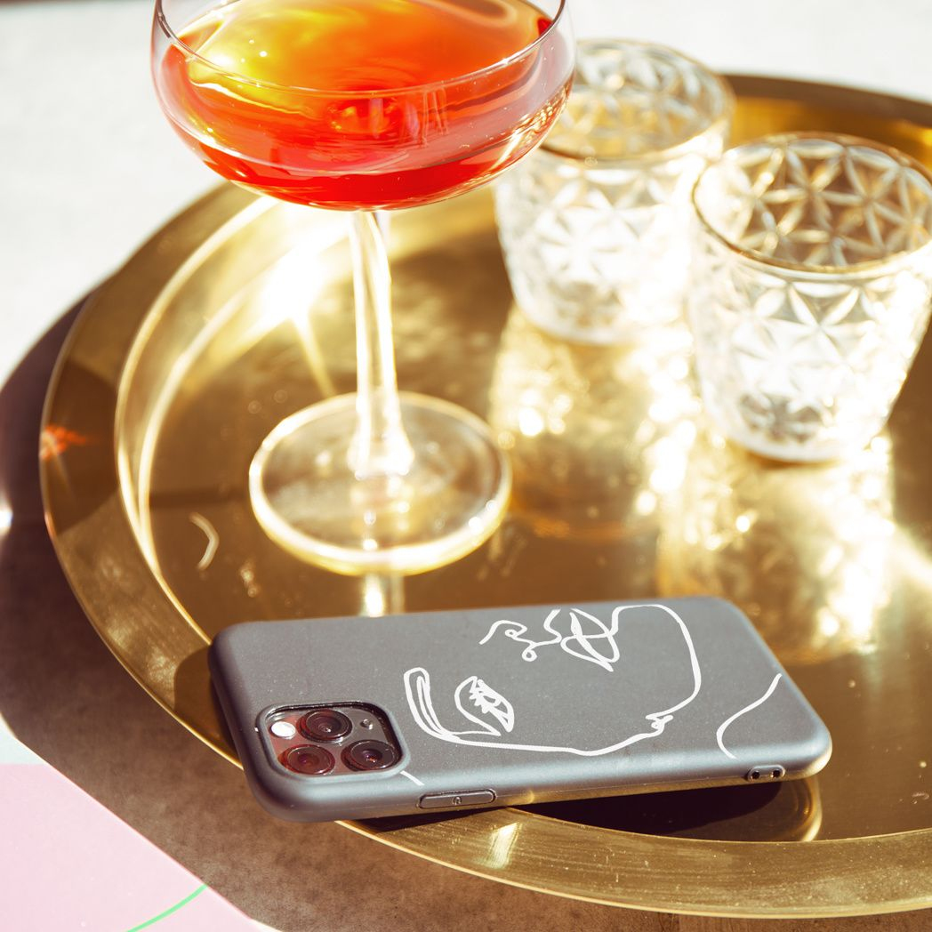 Een cocktail in een glas, twee decoratieve glazen en een telefoonhoesje met een gezichtsontwerp op een gouden dienblad.