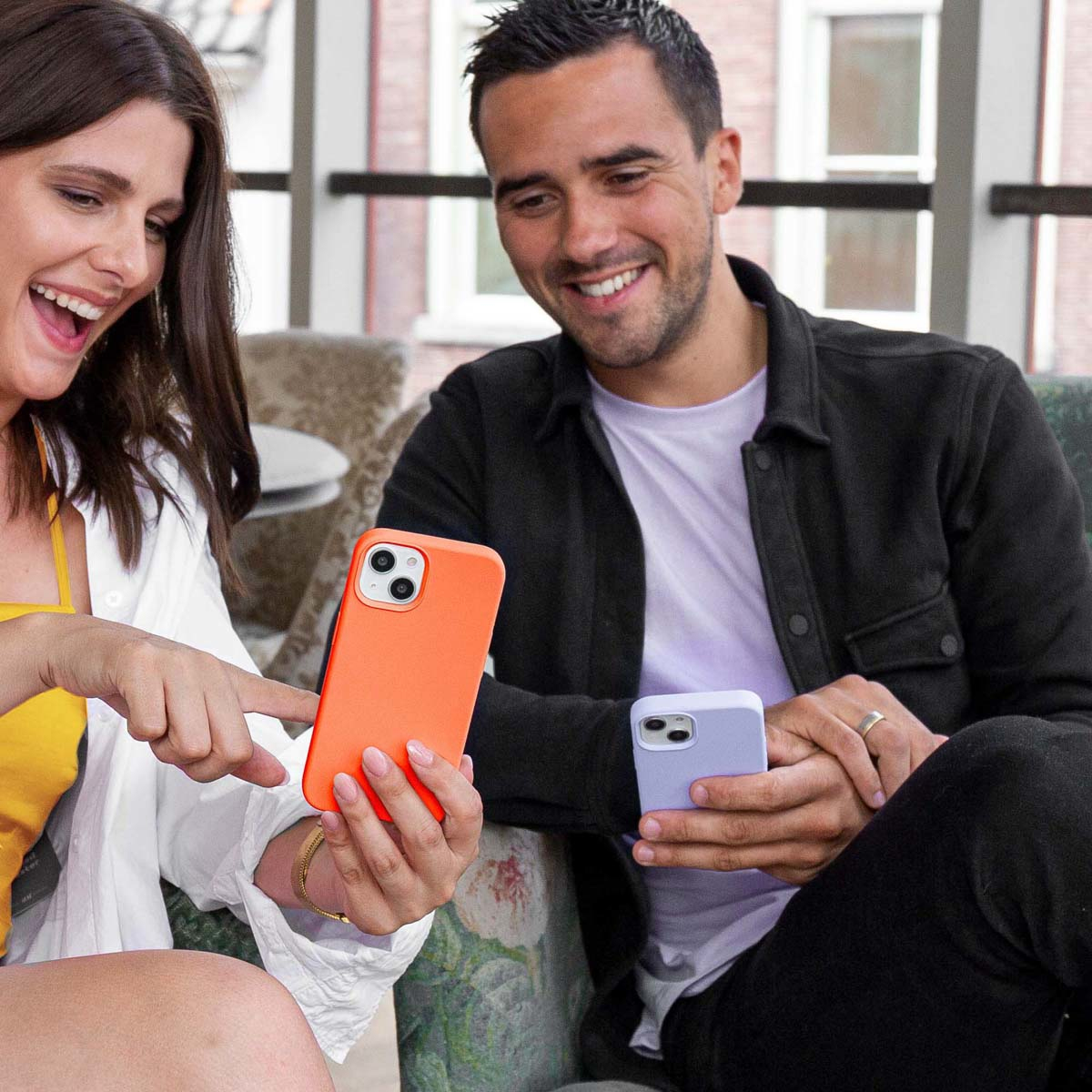 Een man en een vrouw lachen, met telefoons in de hand. De vrouw wijst naar een oranje telefoon. De man houdt een paarse telefoon vast.