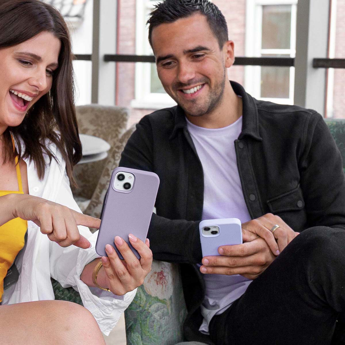 Un homme et une femme sourient, tenant des étuis de téléphone lavande. La femme en montre un du doigt. Ils sont assis dans une pièce avec une fenêtre.