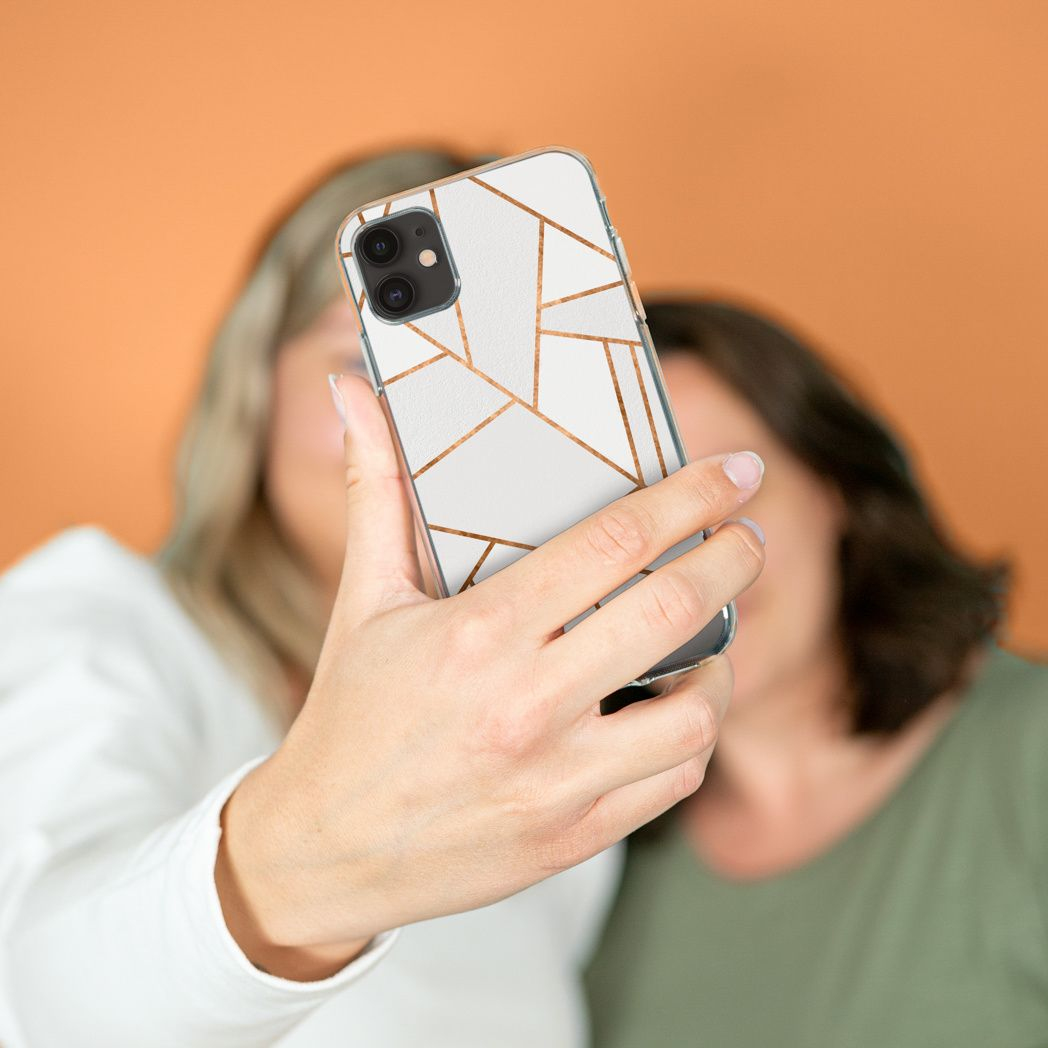 Una persona tiene in mano un telefono, scattando un selfie. La custodia del telefono ha un design geometrico bianco con linee dorate. Altre due persone sono sullo sfondo.