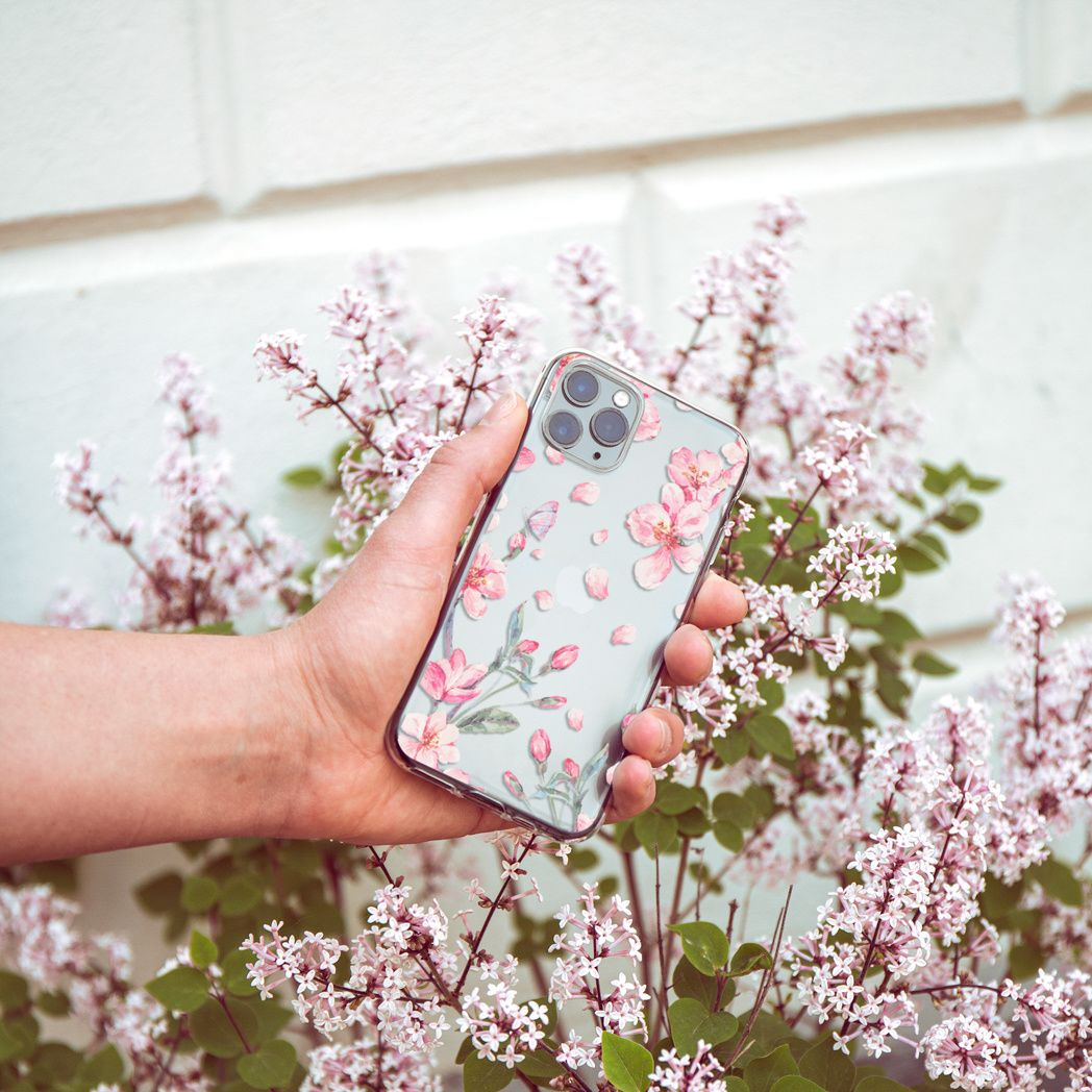 Main tenant un téléphone avec étui floral. Fleurs roses et arbustes de lilas en arrière-plan, contre un mur blanc.