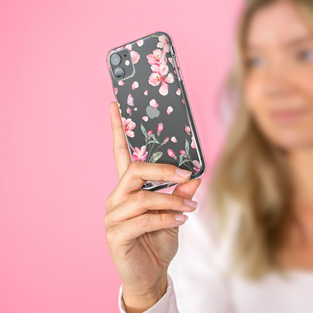 Une main tient un téléphone avec une coque florale. Des fleurs et des pétales roses ornent la coque. Fond rose.