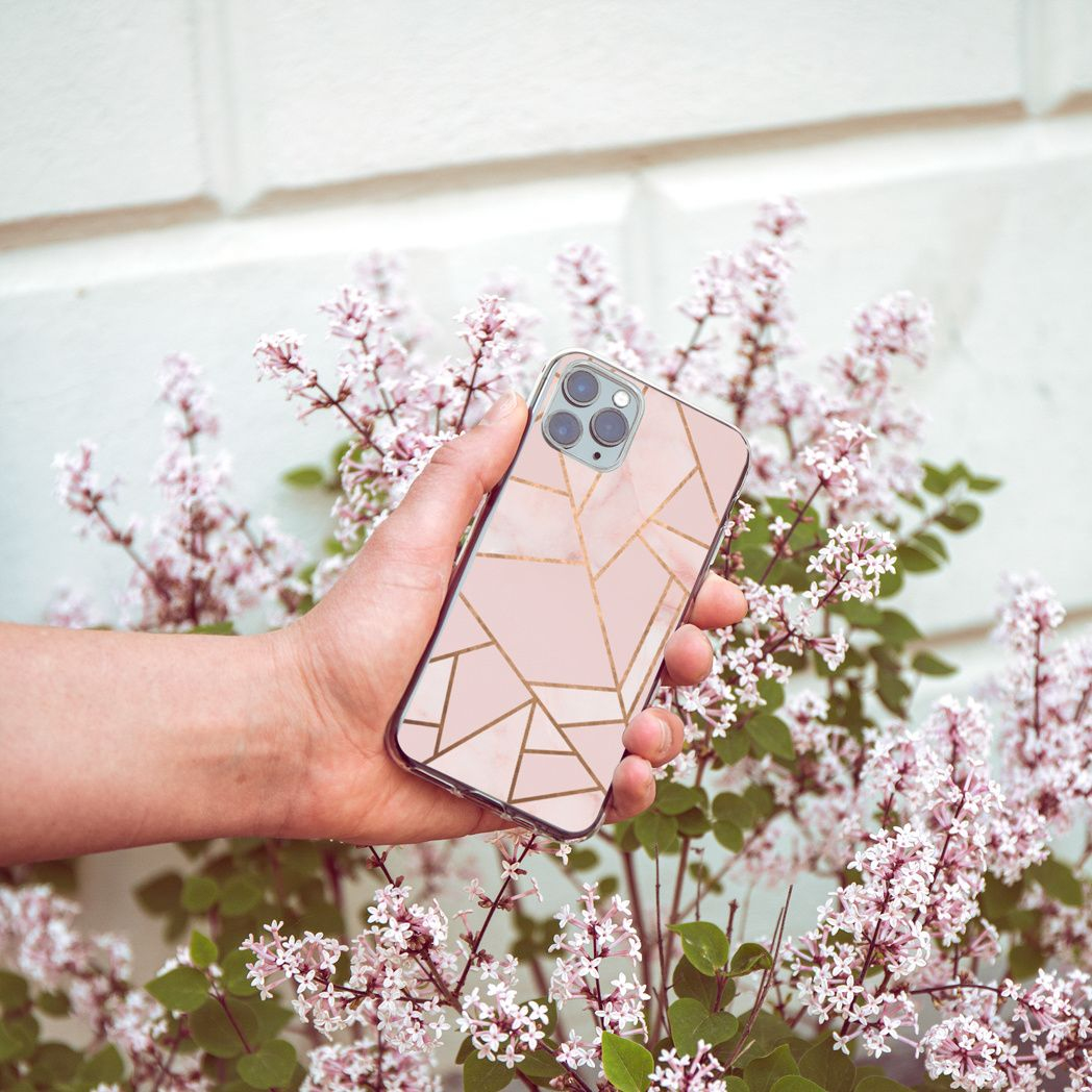 Une main tient un téléphone avec une coque géométrique rose, sur fond de fleurs lilas et de mur blanc.