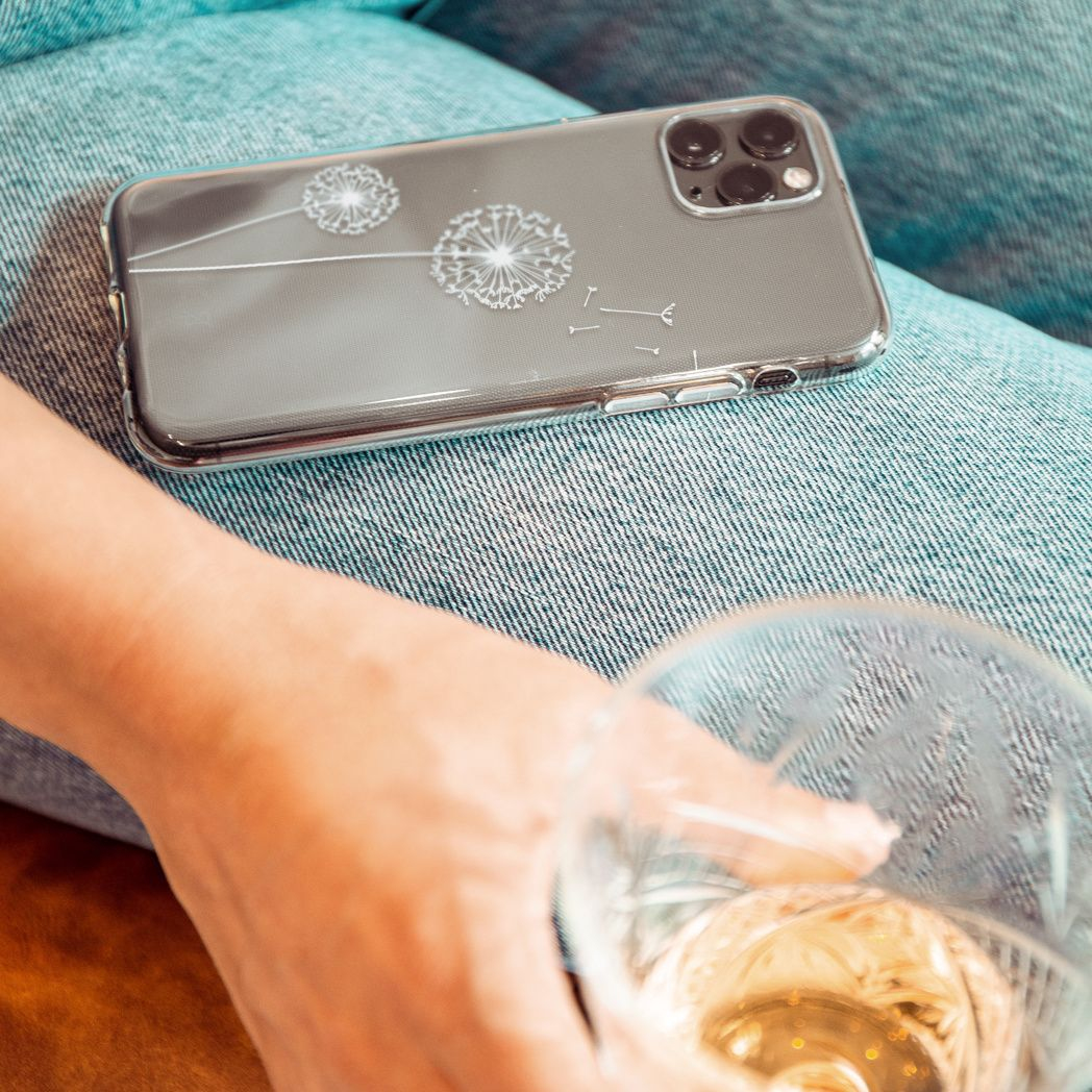Un téléphone avec un étui de pissenlit repose sur du denim. Une main tient un verre de vin. L'arrière-plan est flou.