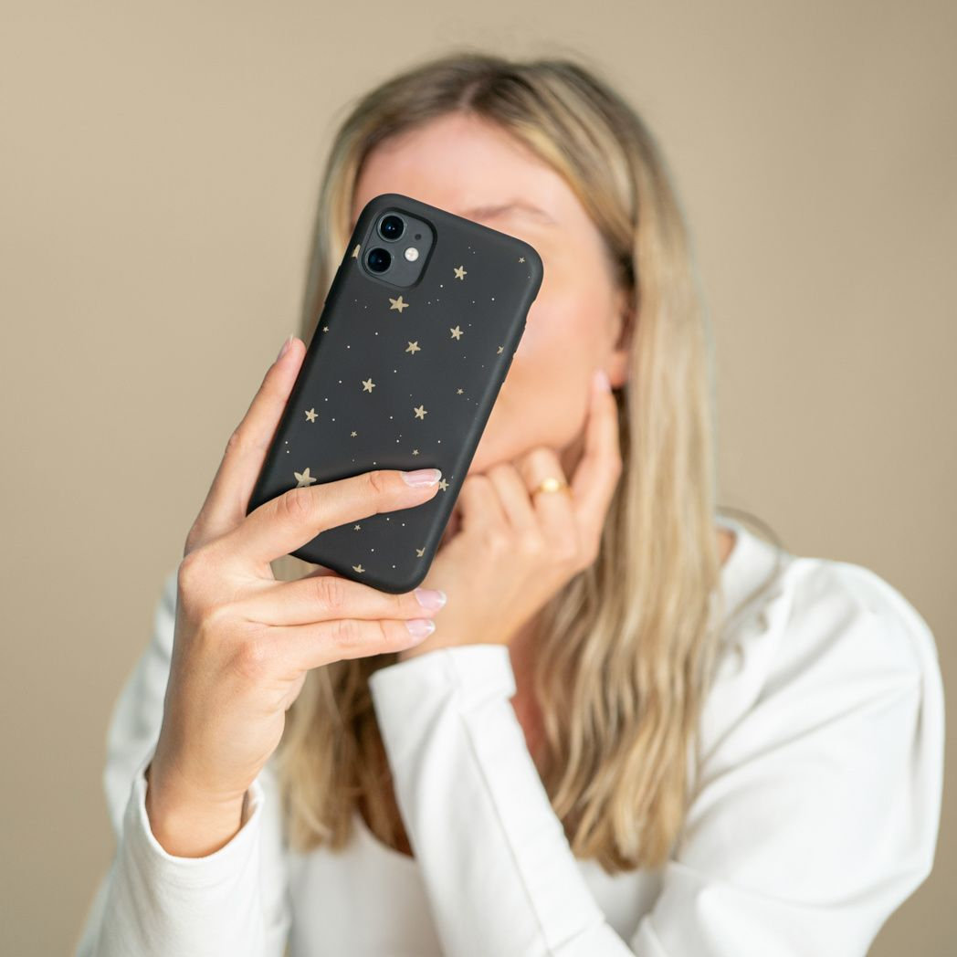 Una donna tiene un telefono nero con stelle dorate vicino al viso. Indossa un top bianco e un anello d'oro. Capelli biondi.
