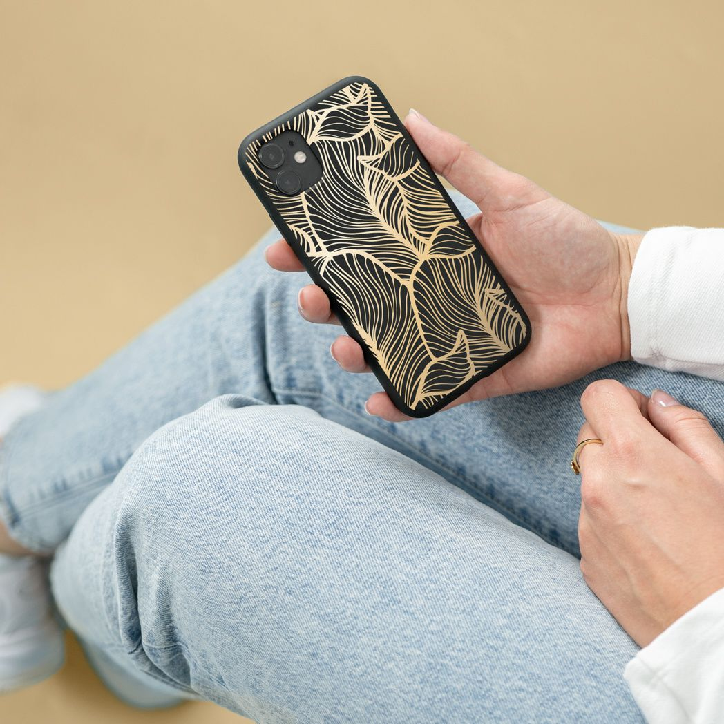 Una persona tiene in mano una custodia nera per telefono con un design a foglie dorate. Indossa jeans e una camicia bianca. È visibile un anello d'oro.