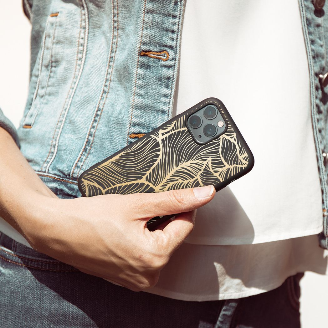 Una persona tiene in mano un telefono con una custodia con un design a foglie nere e oro. Indossa una giacca di jeans e una camicia bianca.