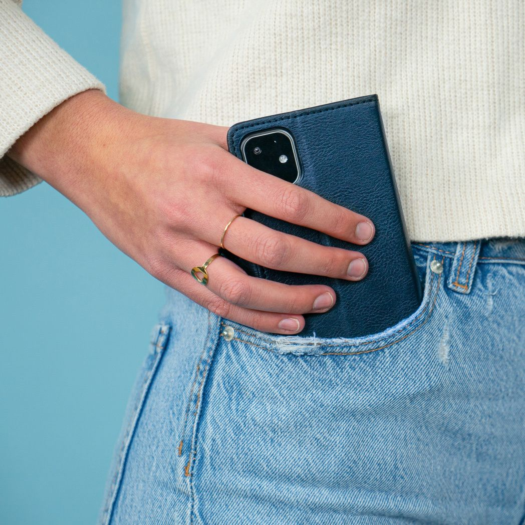 Une personne portant un pull blanc et un jean tient un étui de téléphone bleu dans sa poche. Une bague en or orne son doigt.