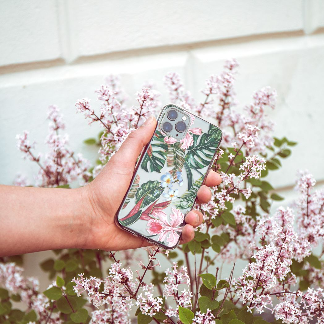 Hand hält Handy mit floralem Case. Blumen im Hintergrund, darunter weiße, rosa und grüne Blätter. Weiße Wand.