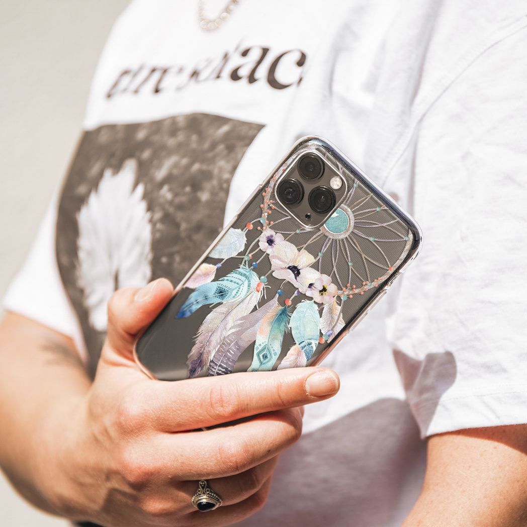 Una persona tiene in mano un telefono con una custodia con un design acchiappasogni. La custodia ha piume e fiori. La persona indossa una camicia bianca.