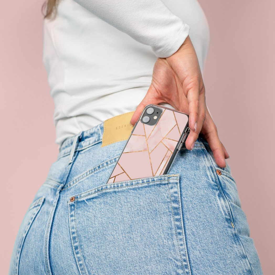 Una persona infila un telefono con una custodia dal design geometrico rosa e oro in una tasca di jeans blu. Si vede una camicia bianca a maniche lunghe.