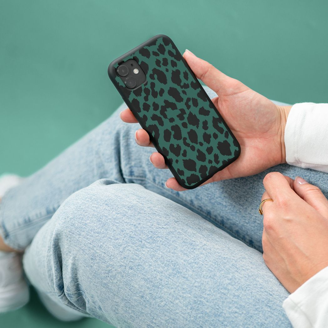 Una persona tiene in mano un telefono con una custodia verde con stampa leopardata. Indossa jeans e una camicia bianca. C'è un anello d'oro al dito.