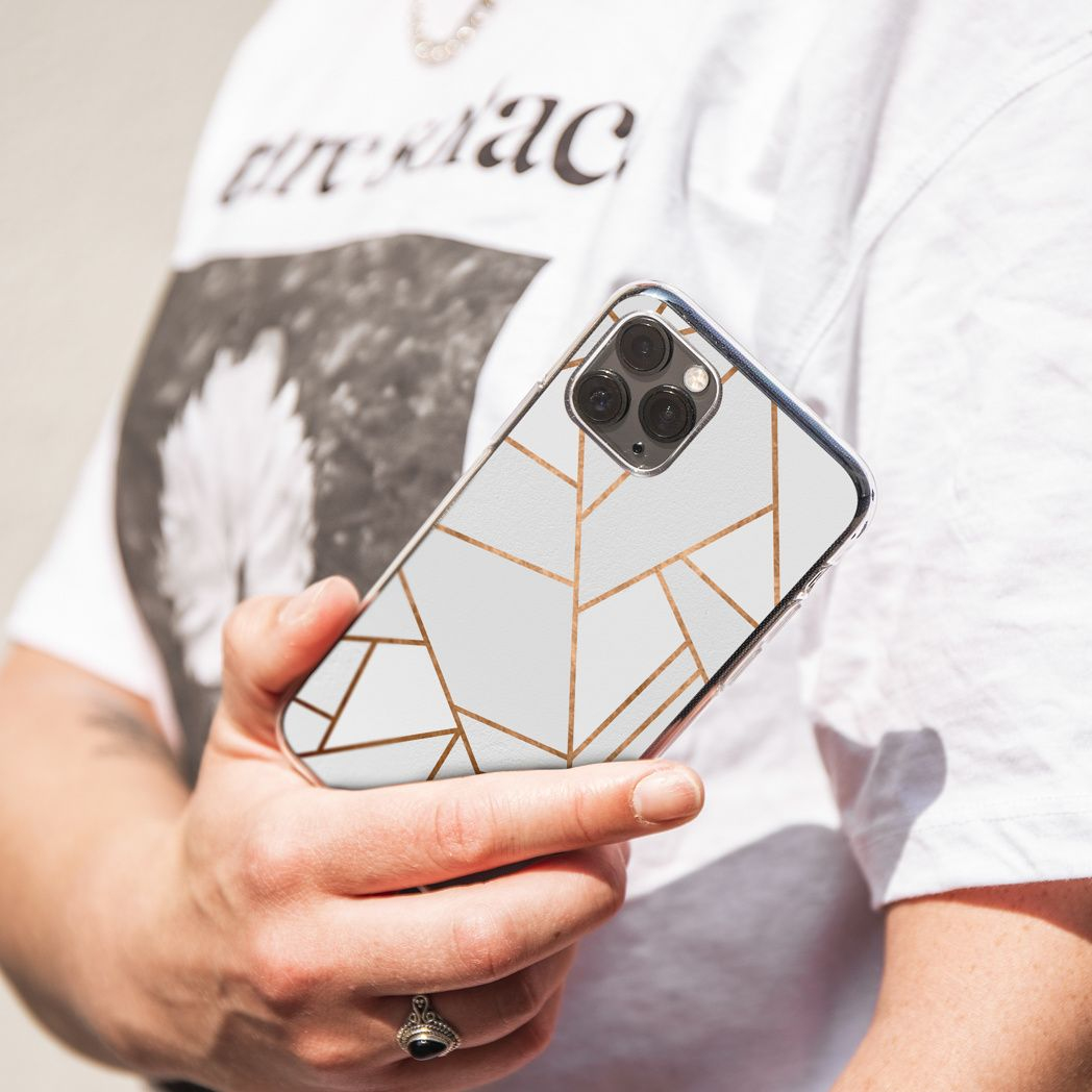 Een persoon houdt een telefoon vast met een witte hoes en gouden geometrische lijnen. Ze dragen een wit shirt en een ring.