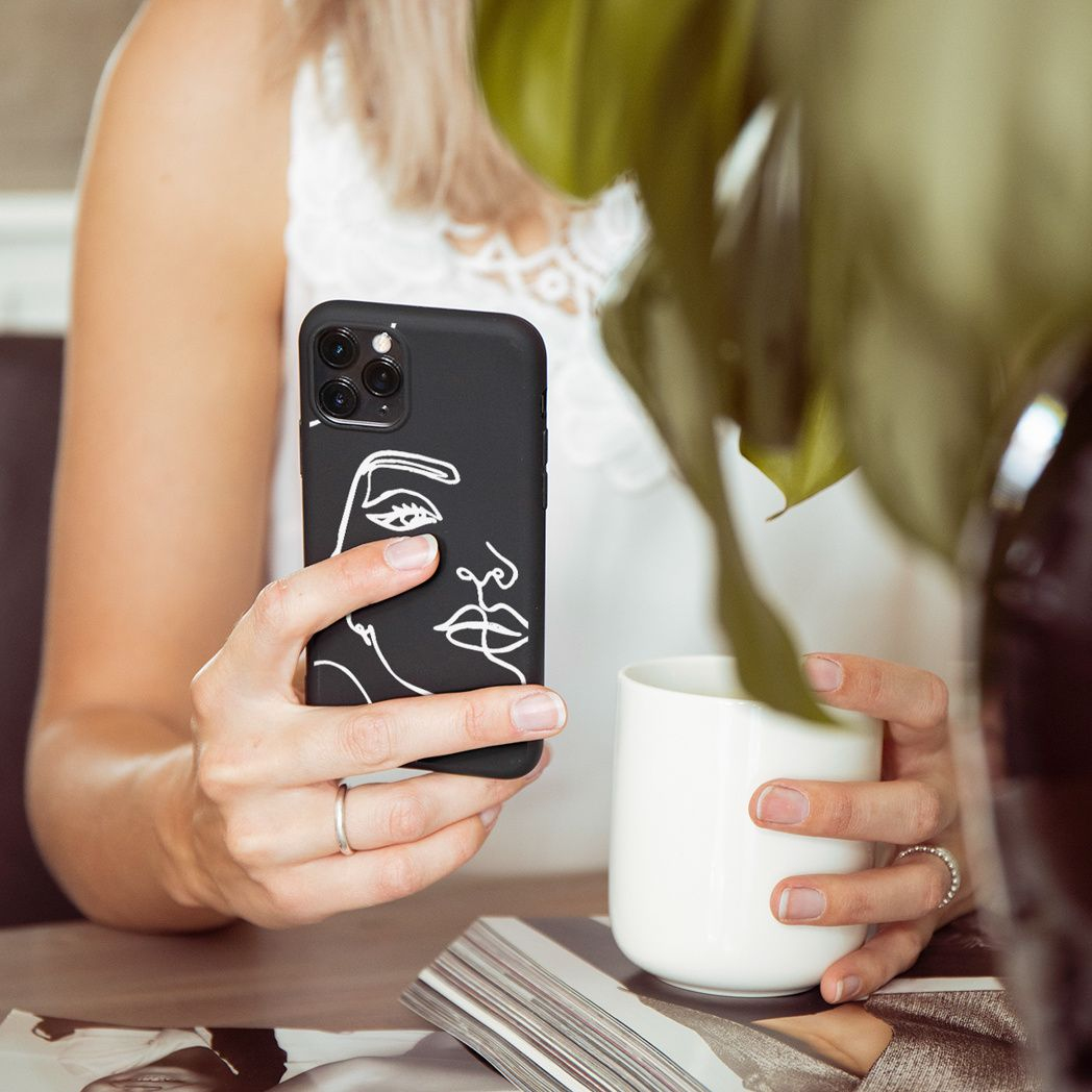 Una donna tiene in mano un telefono nero con un disegno di volto e una tazza bianca. Indossa un top bianco e ha anelli alle dita.