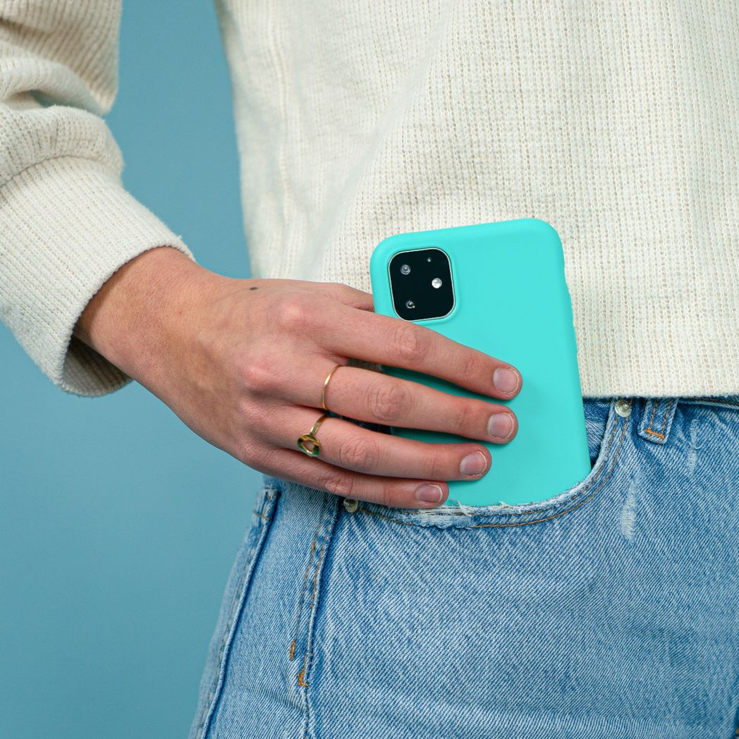 Een persoon houdt een turquoise telefoon in zijn zak. Ze draagt een witte trui en een spijkerbroek. Gouden ringen sieren hun vingers.