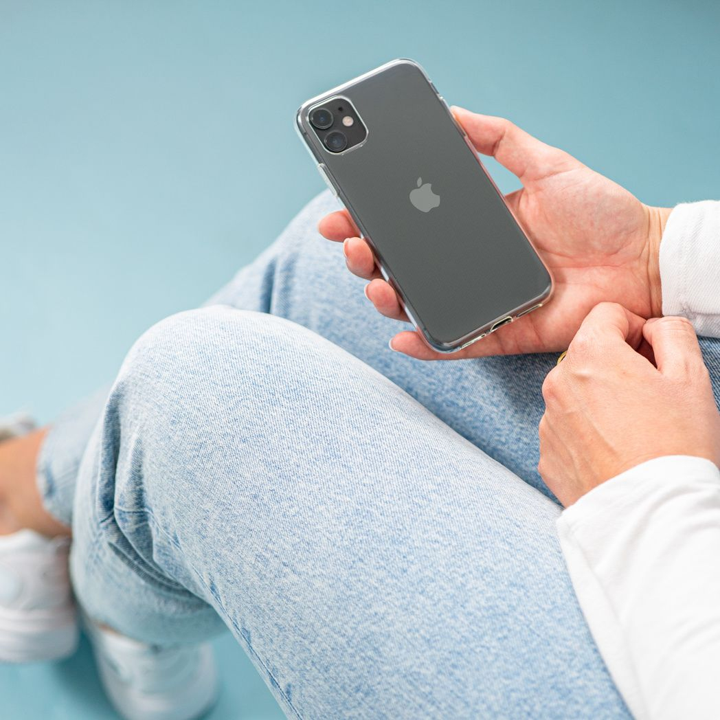 Una persona sostiene un iPhone gris en una funda transparente. Lleva vaqueros azul claro y una camisa blanca. El fondo es azul claro.