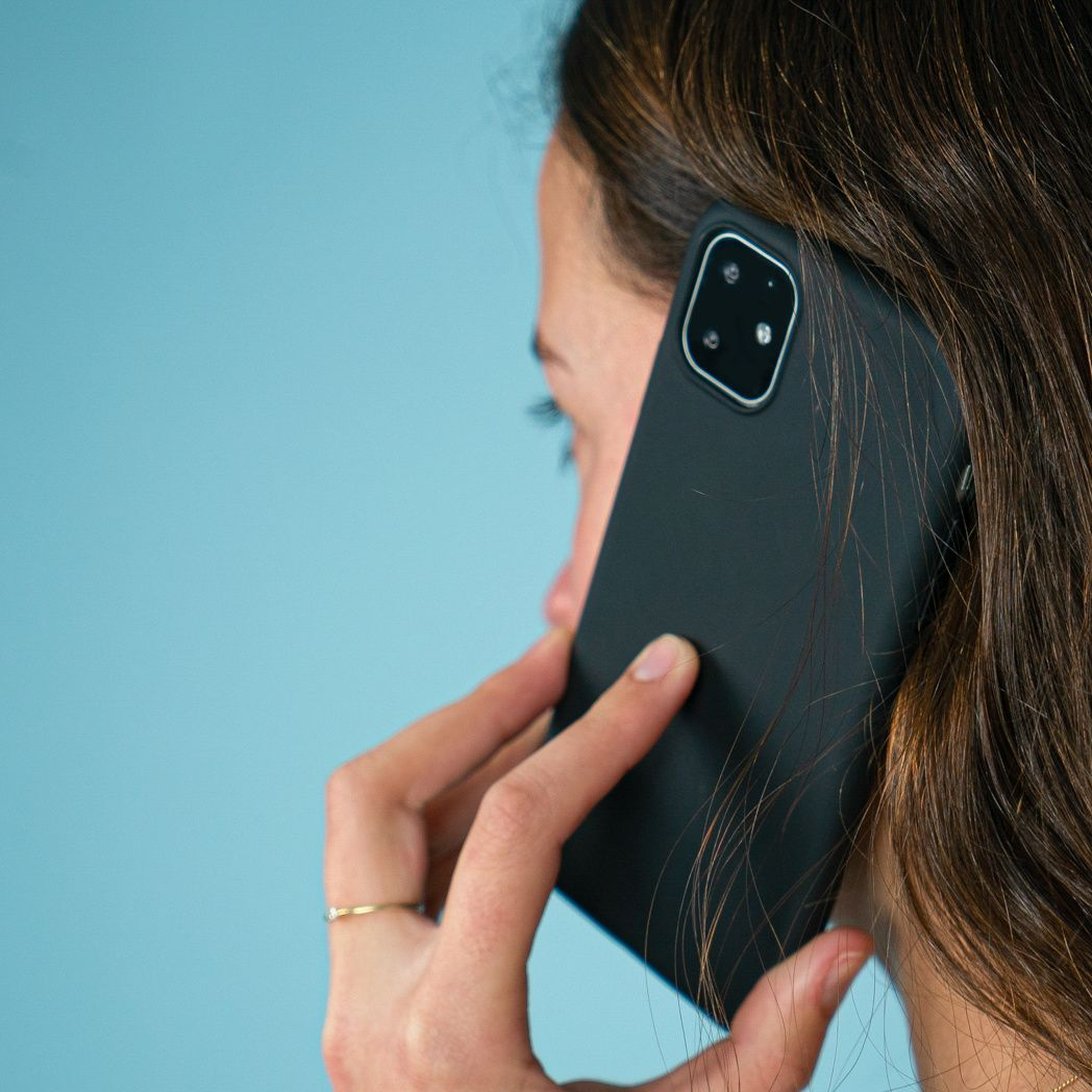 Een vrouw houdt een zwarte telefoon aan haar oor. Ze heeft bruin haar en een gouden ring. De achtergrond is lichtblauw.