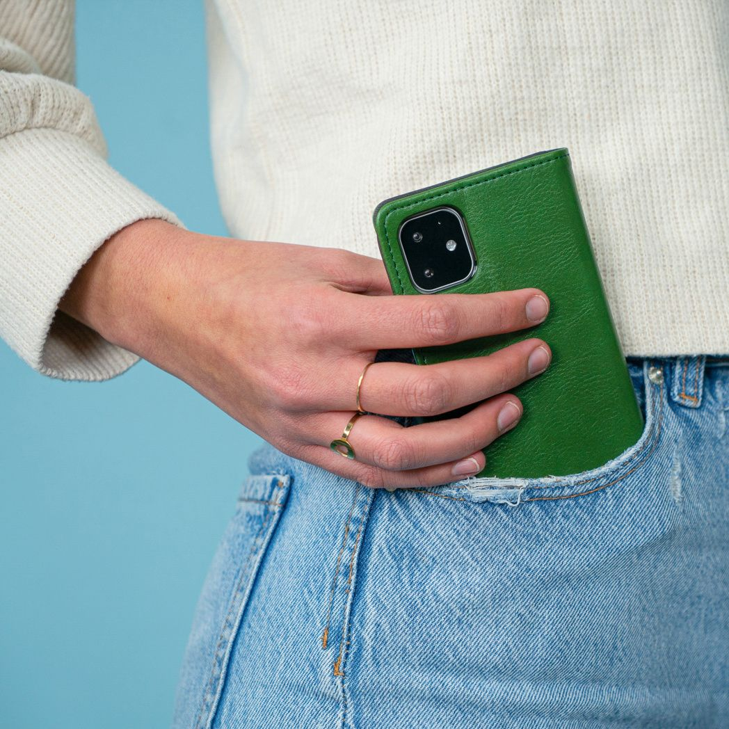 Una mano tiene una custodia verde per telefono, mettendola in una tasca dei jeans. La persona indossa un maglione bianco e un anello d'oro. Sfondo blu.