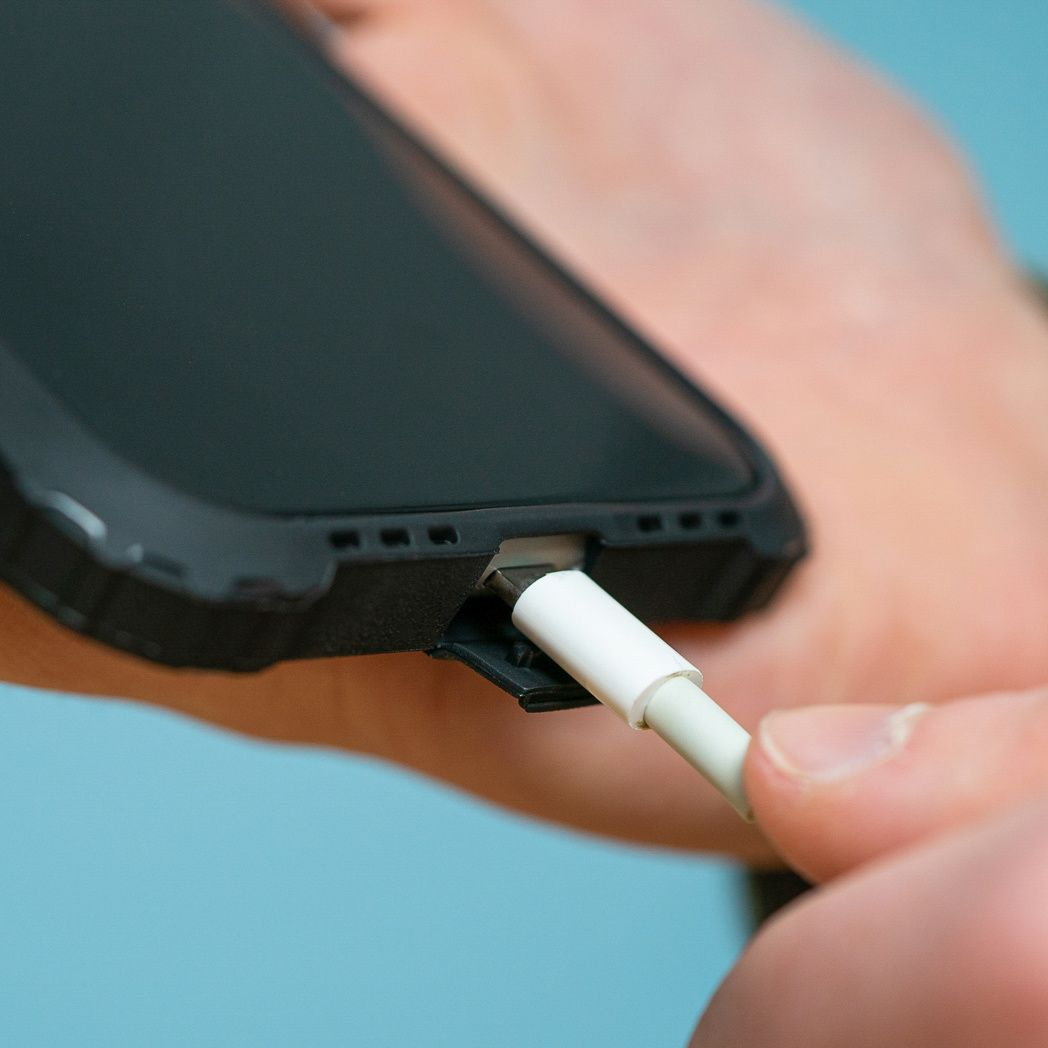 Een hand steekt een witte oplaadkabel in een zwarte telefoon. De telefoon heeft een zwarte hoes en de achtergrond is lichtblauw.