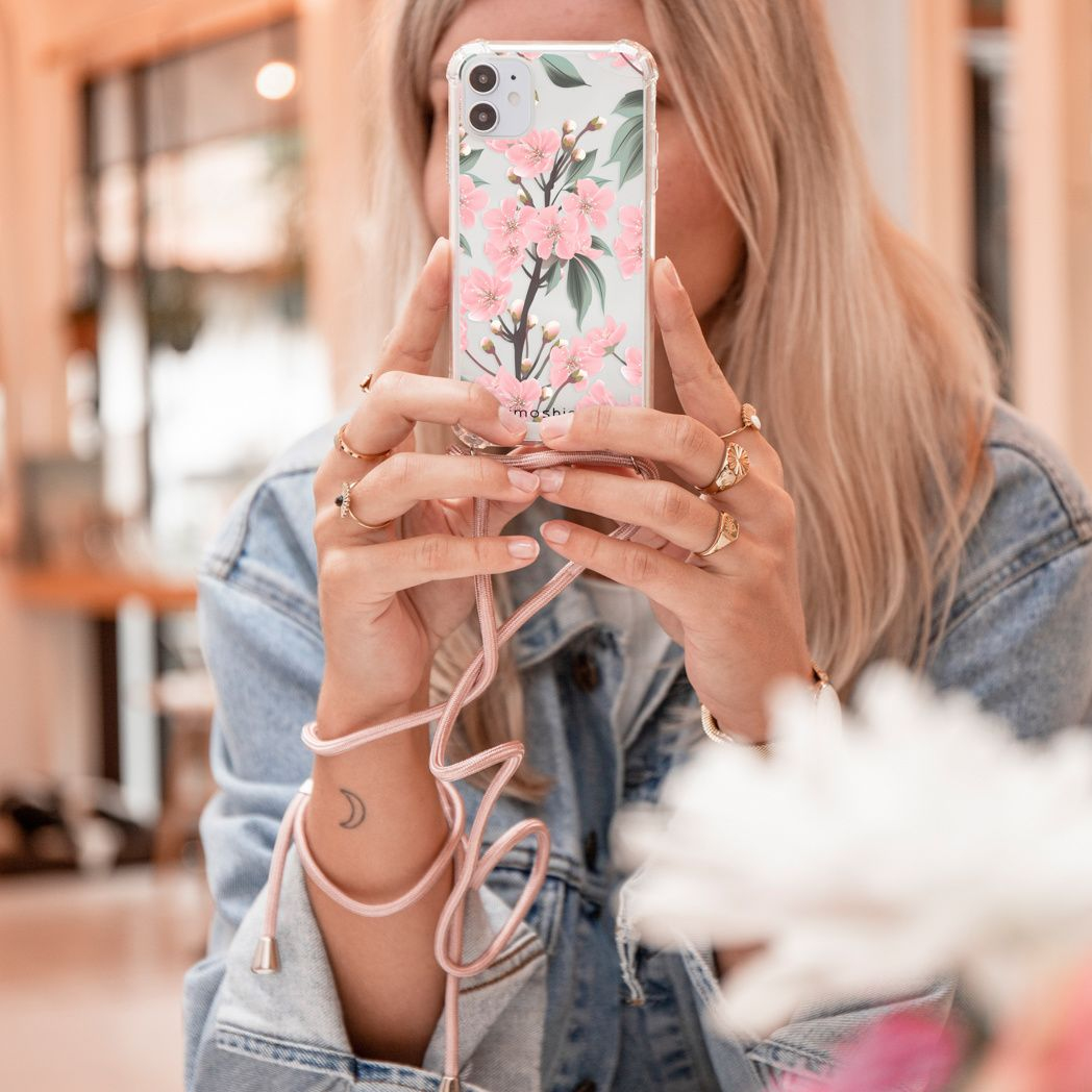 Een vrouw houdt een telefoon met een bloemenhoes vast. Ze draagt een spijkerjasje en ringen. Een roze koord hangt van de telefoon.