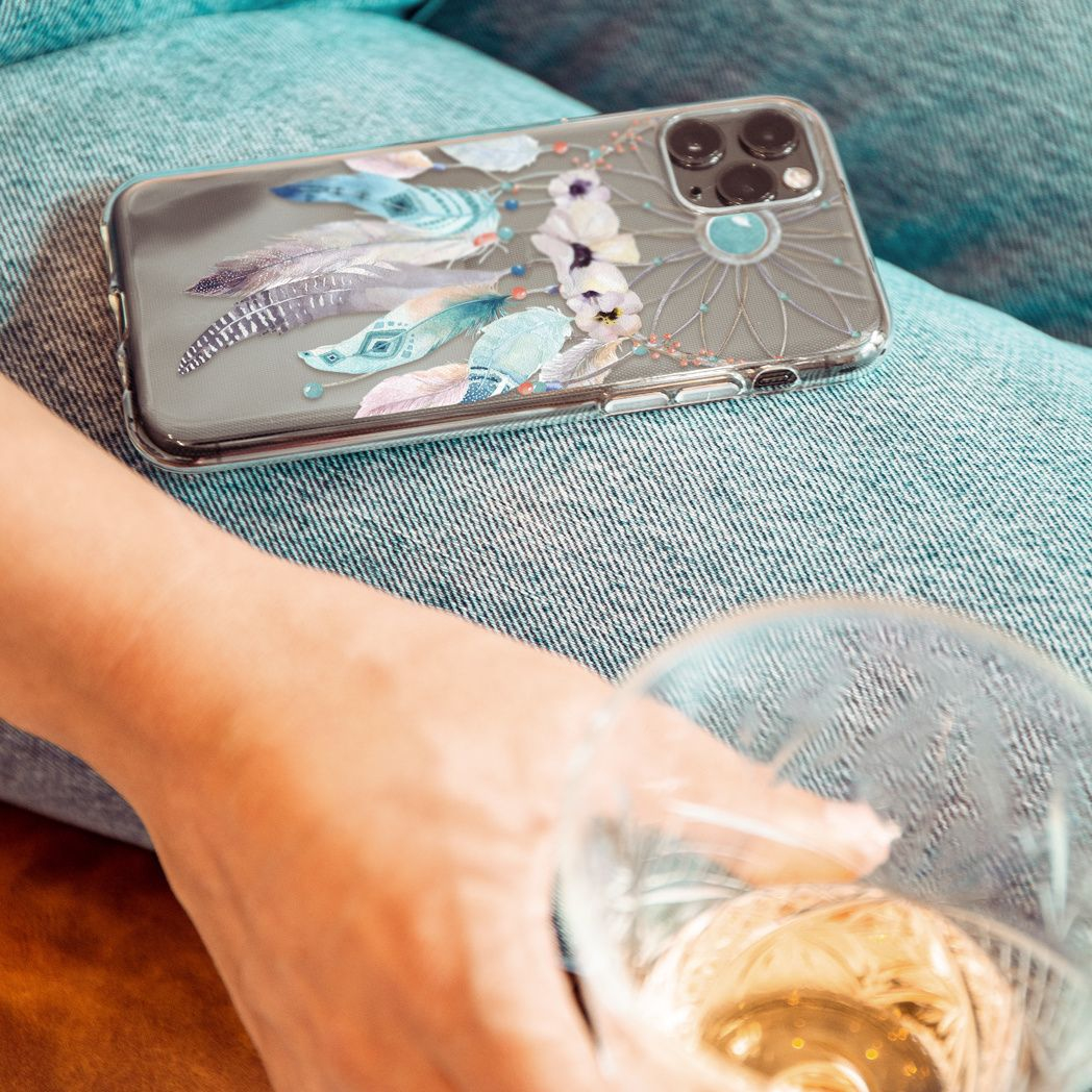 Un téléphone avec une coque attrape-rêves repose sur un jean. Une main tient un verre de vin avec un liquide doré. Flou.