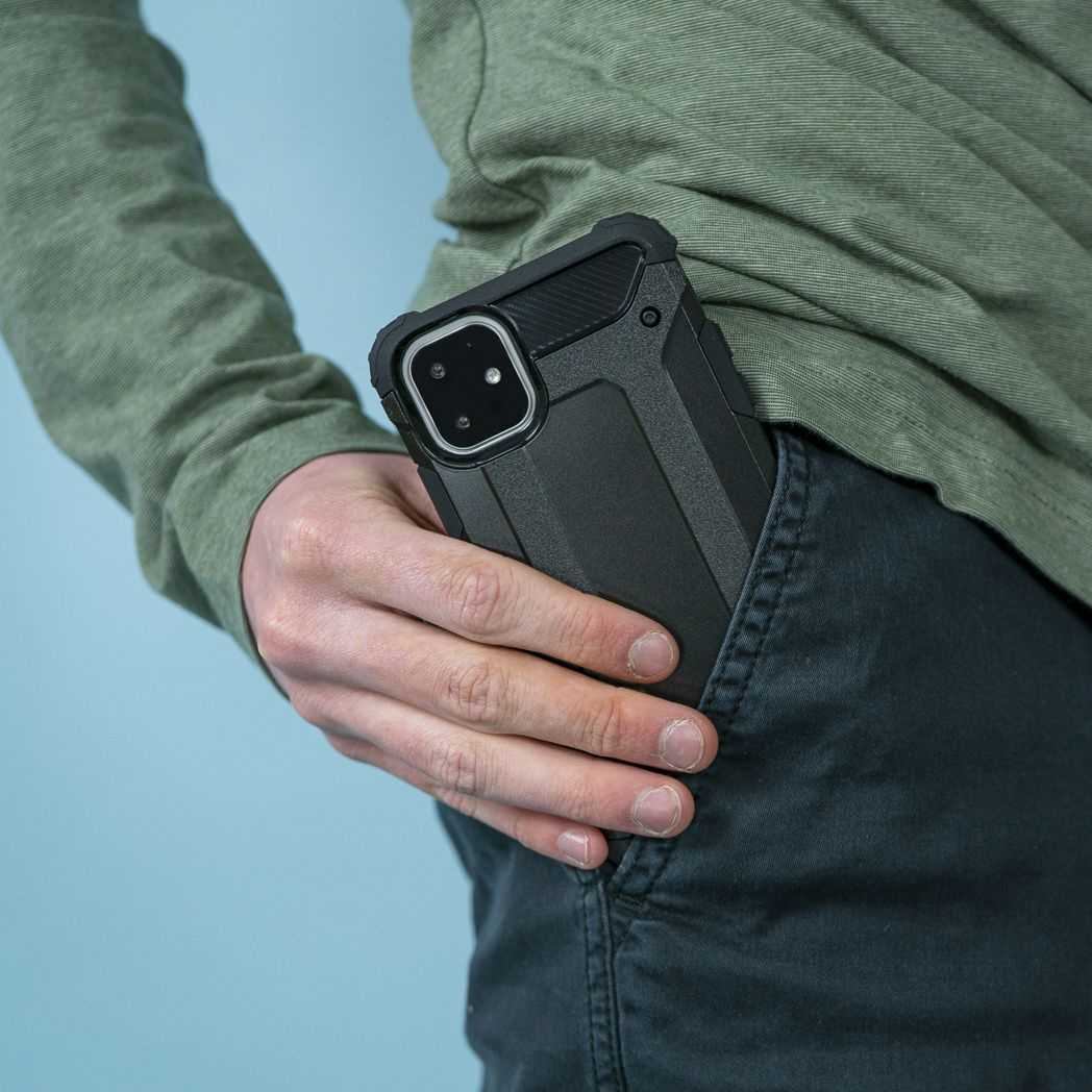 Una persona tiene in tasca un telefono nero. La persona indossa una camicia verde e pantaloni scuri su uno sfondo azzurro.