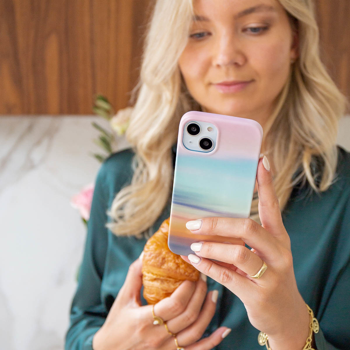 Blonde vrouw die een telefoon met een kleurrijke hoes en een croissant vasthoudt. Ze draagt een groen shirt en gouden sieraden.
