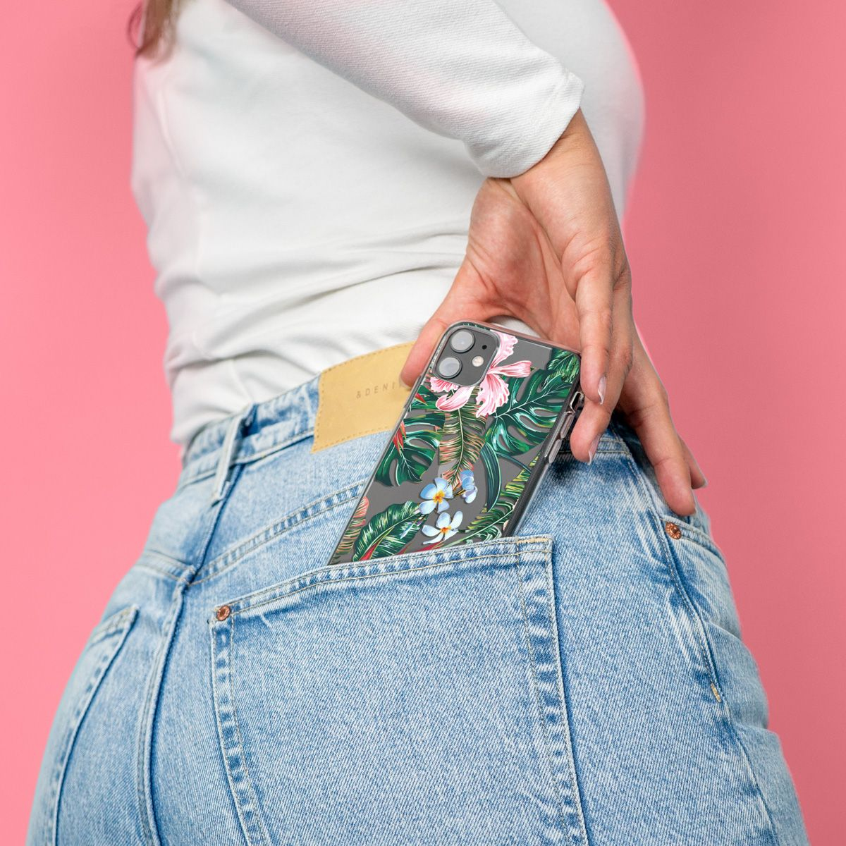 Une personne met un téléphone avec un étui floral dans une poche de jean. Le fond est rose. L'étui a des fleurs et des feuilles.