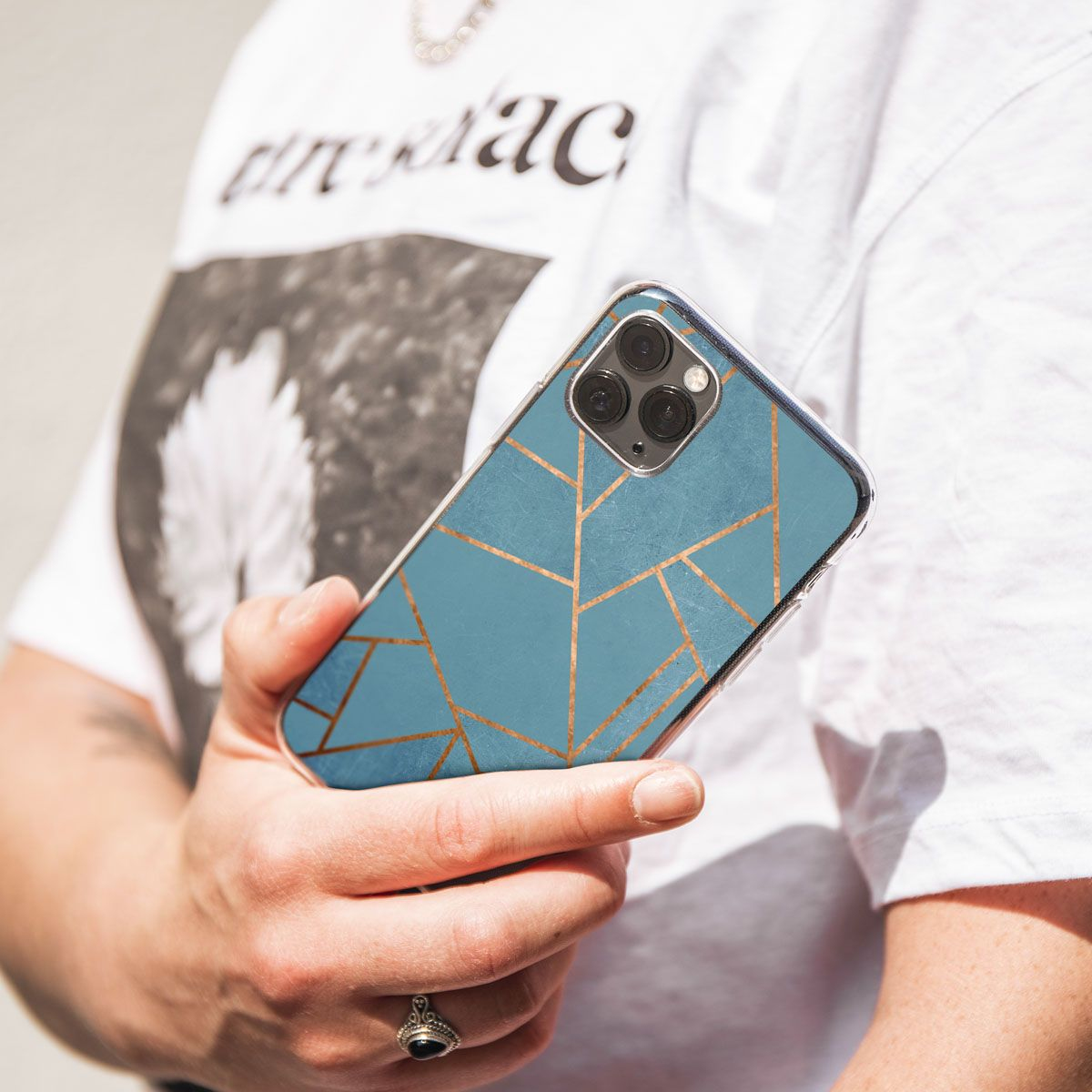 Eine Person hält ein Telefon mit einer blau-goldenen geometrischen Hülle. Sie trägt ein weißes T-Shirt und einen Ring.