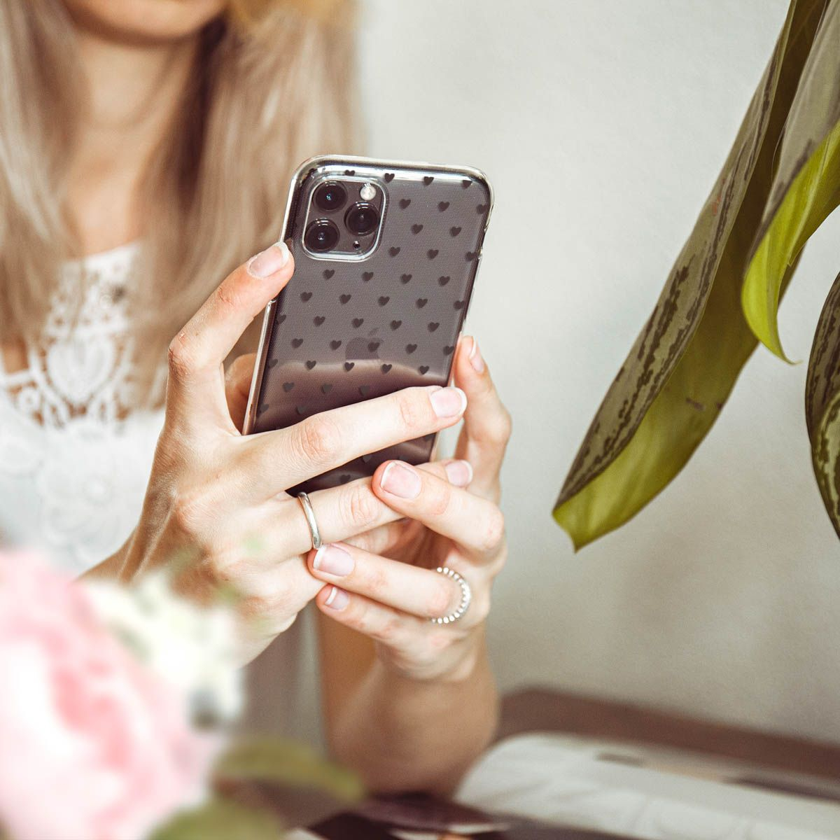 Donna che tiene in mano un telefono con una custodia a forma di cuore. Anelli alle dita, pianta sullo sfondo.