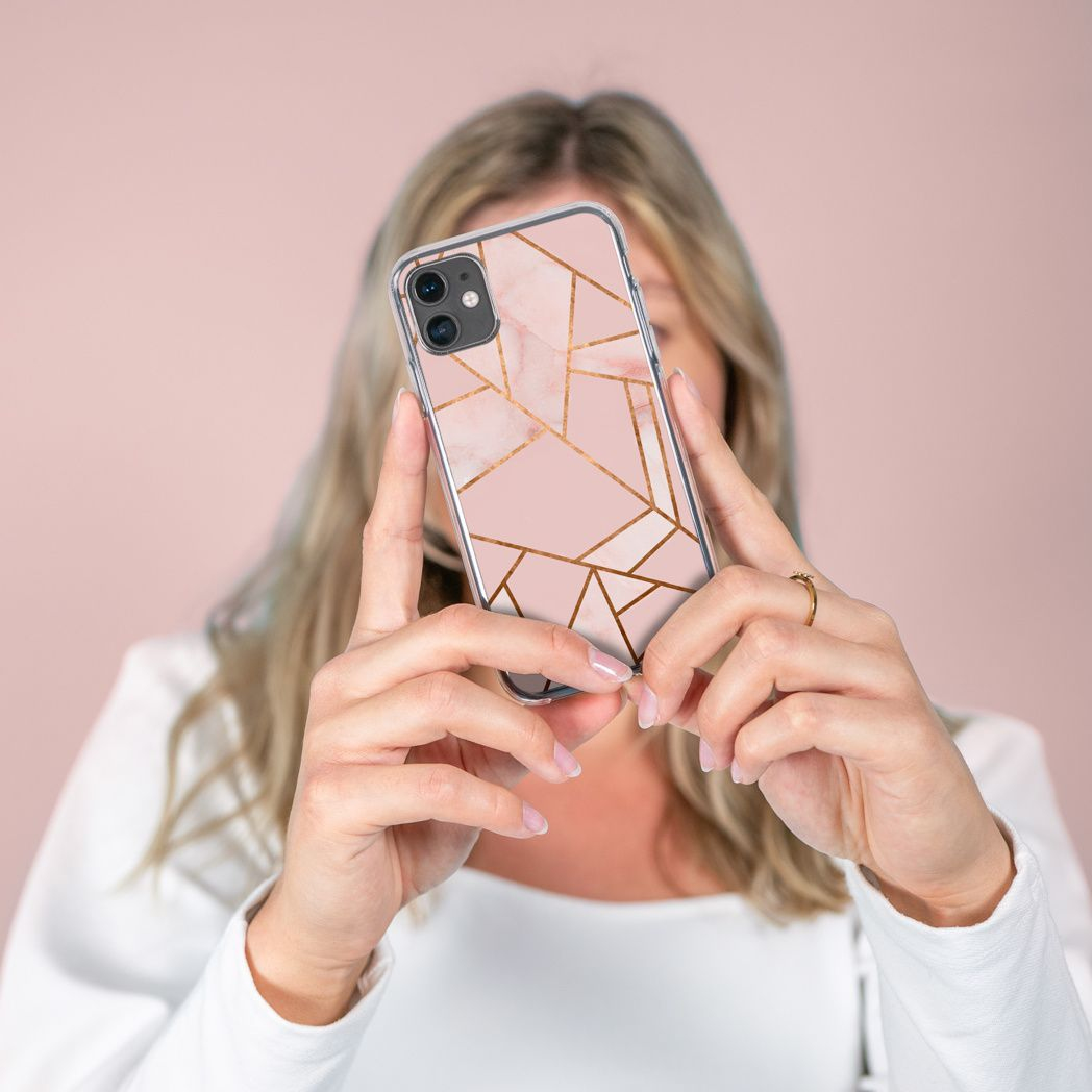 Une femme tient un téléphone avec un étui géométrique rose. Le fond est rose clair. Elle porte une bague et une chemise blanche.