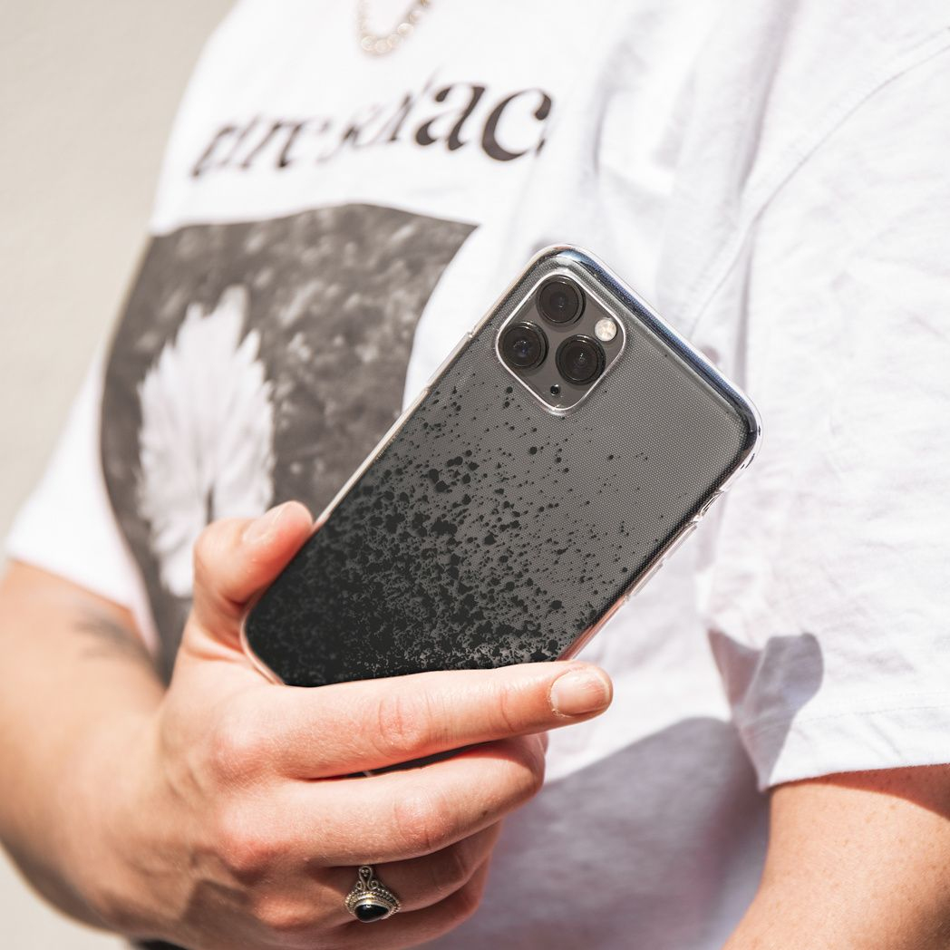 Eine Person hält ein schwarzes Smartphone in einer durchsichtigen Hülle. Die Person trägt ein weißes T-Shirt und einen Silberring. Der Hintergrund ist verschwommen.